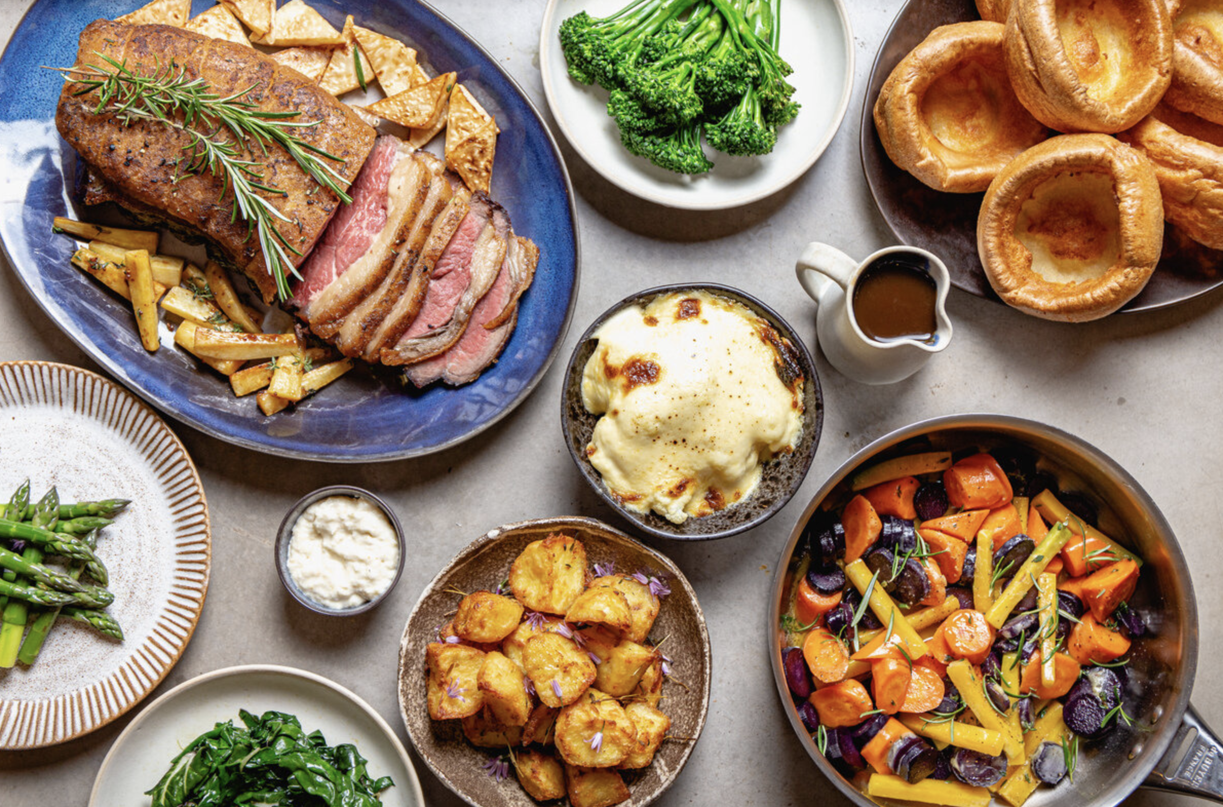 A spread of Sunday Lunch dishes including roast beef, greens, cauliflower cheese and Yorkshire puddings on a white tablecloth