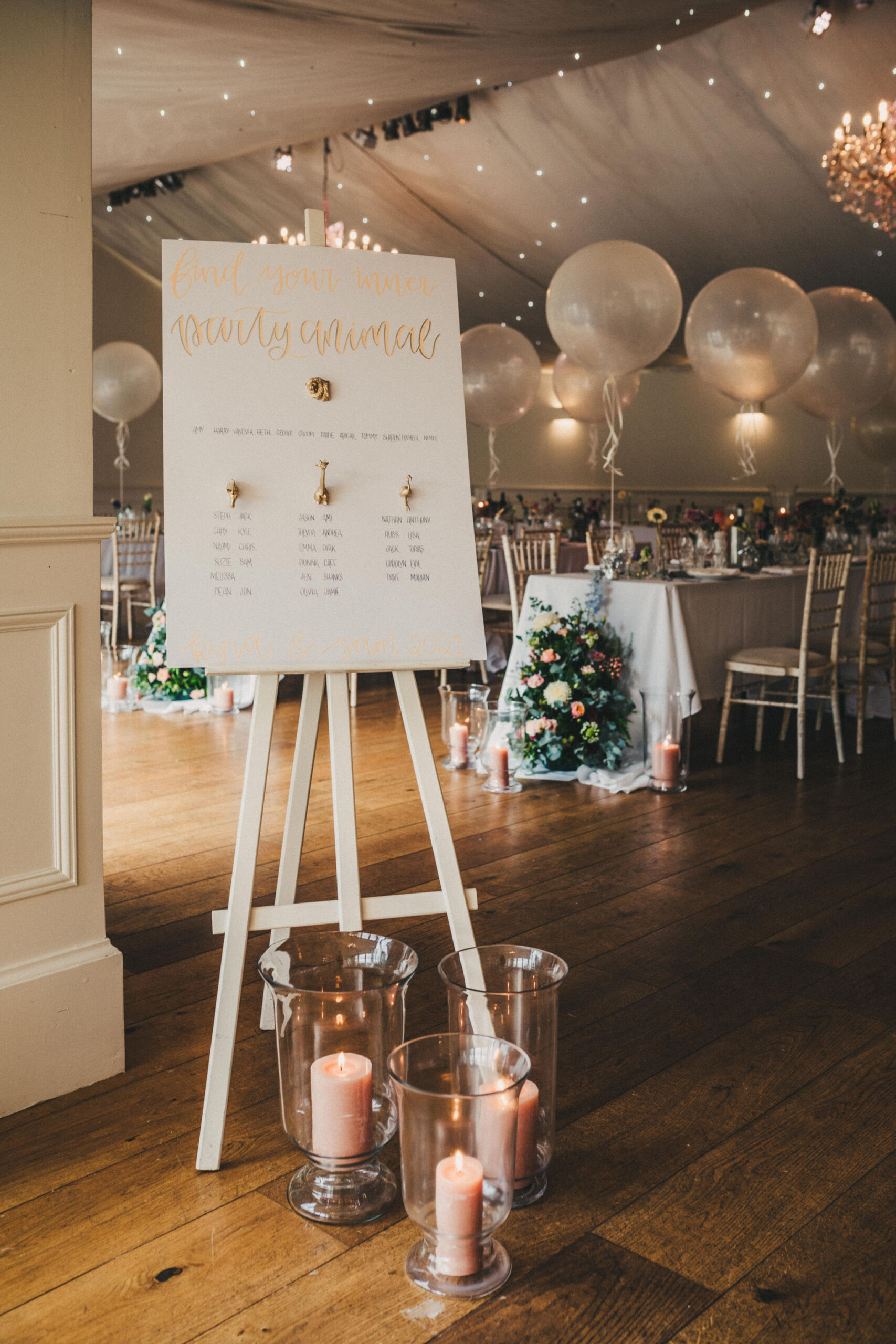 A wedding table plan is propped up on a white easel with tables set up for a wedding breakfast can be seen in the background