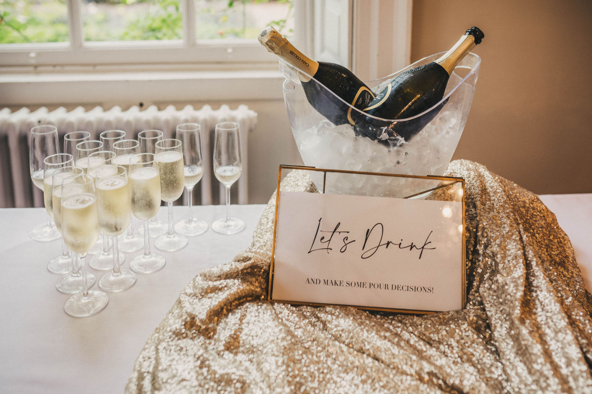 Two bottles of champagne sit in a clear perspex ice bucket with a sign in front of them with the words let's drink