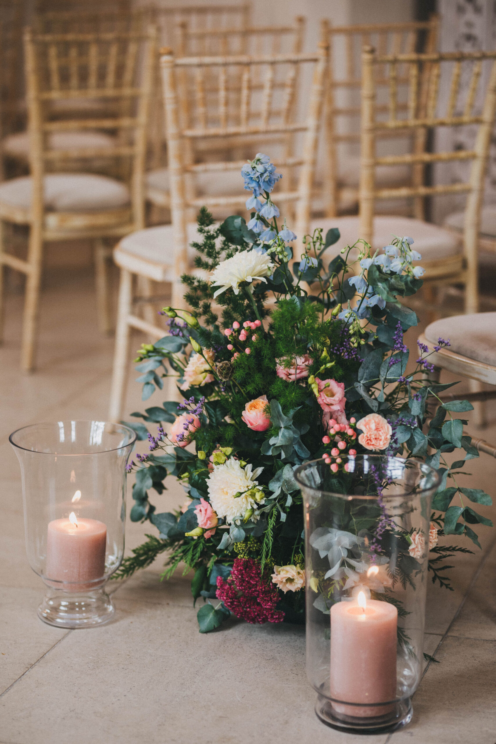 A floral pew end is arranged on the floor of a wedding aisle with pink candles in hurricane vases next to it