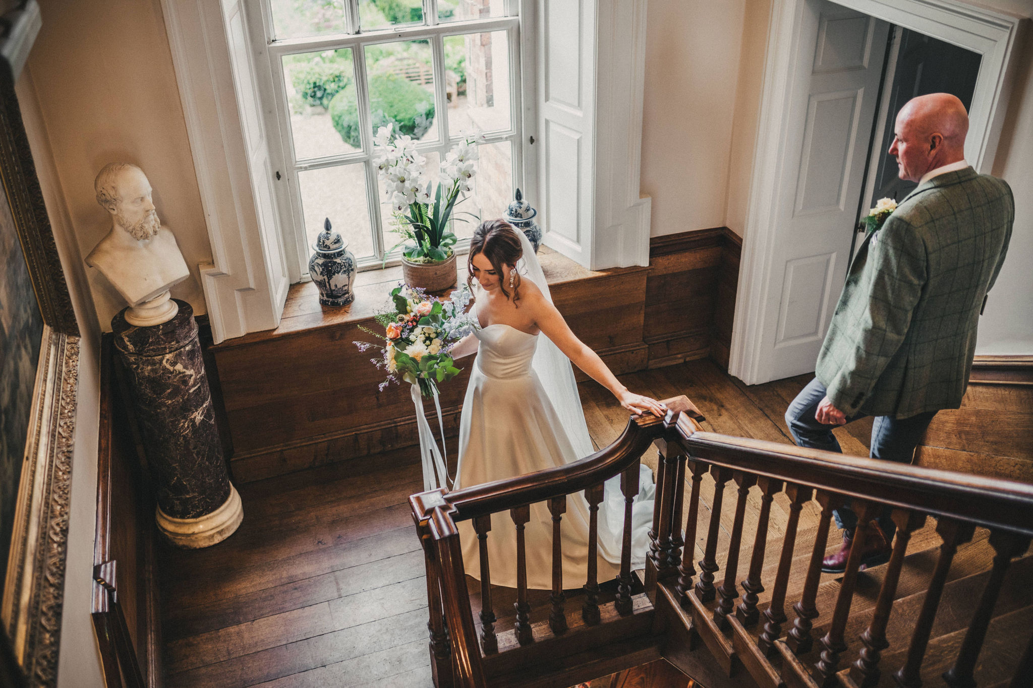 A bride descends an ornate wooden antique staircase holding a pastel coloured bouquet of flowers