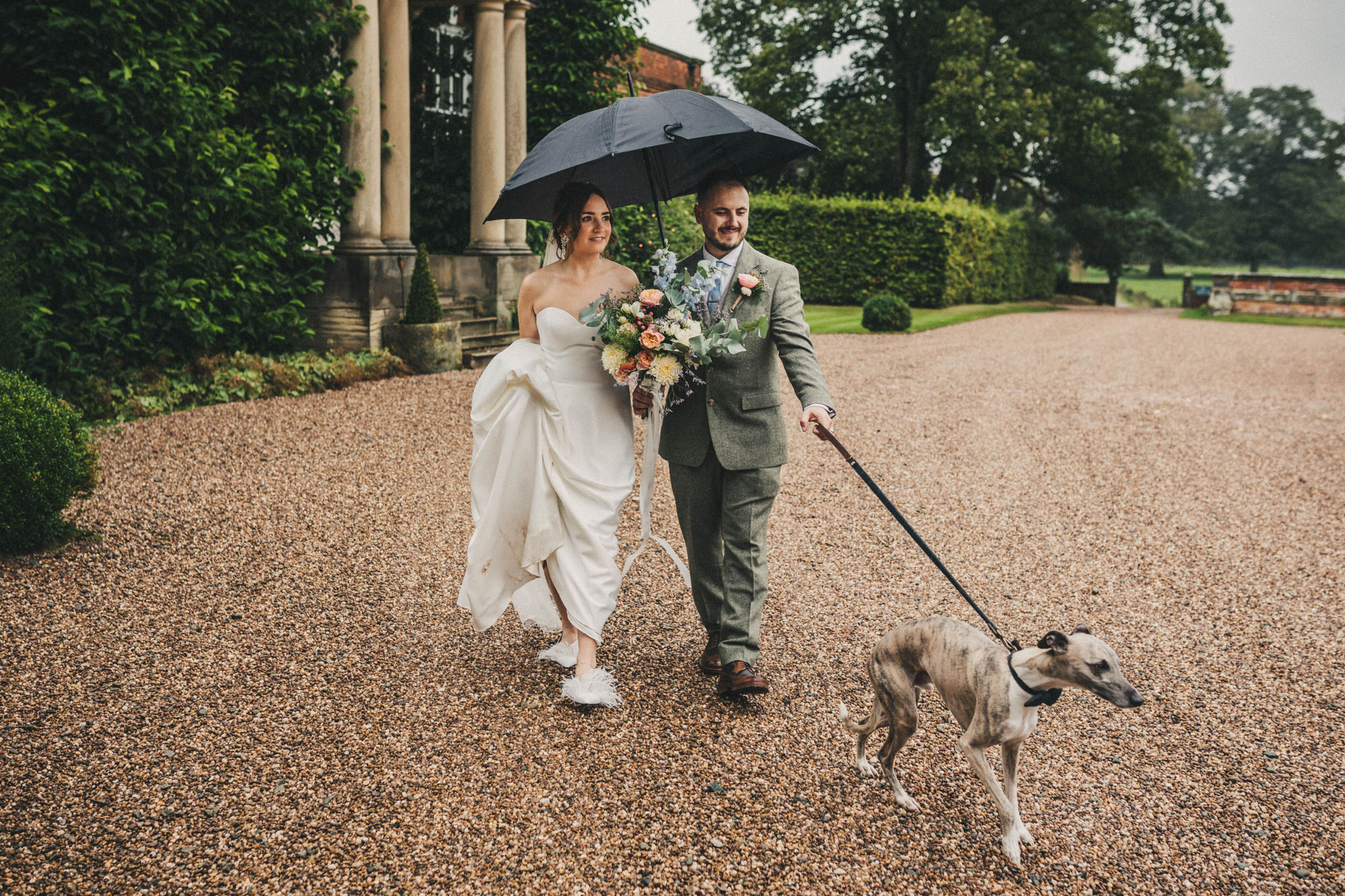A bride and groom parade outside the front of a Georgian Manor House with a gravel drive whilst holding a black umbrella and a grey whippet on a black lead