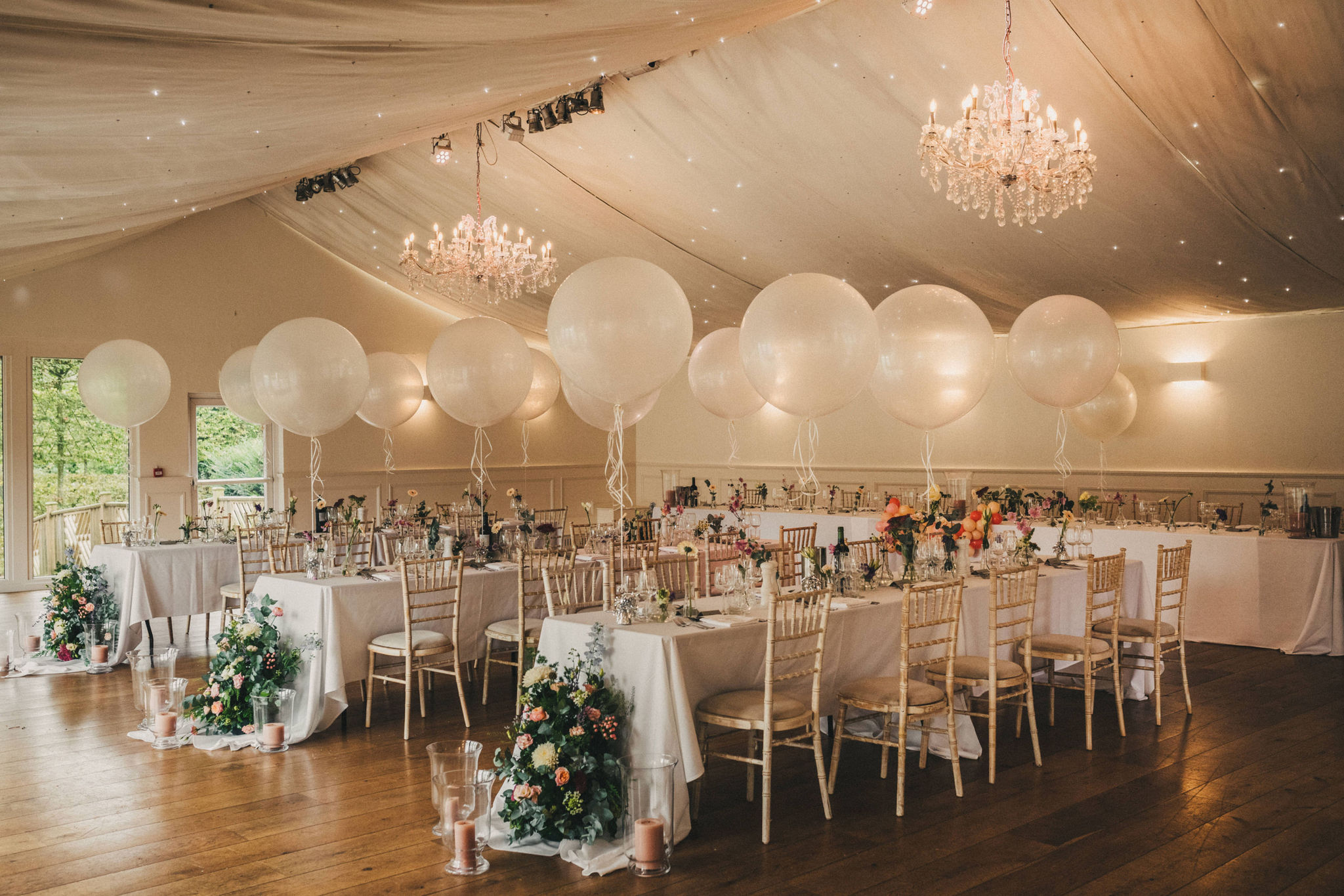 A marquee set up for a country themed wedding with trestle tables covered in white tablecloths and multiple flower arrangements with large white balloons tied to the back of the chairs
