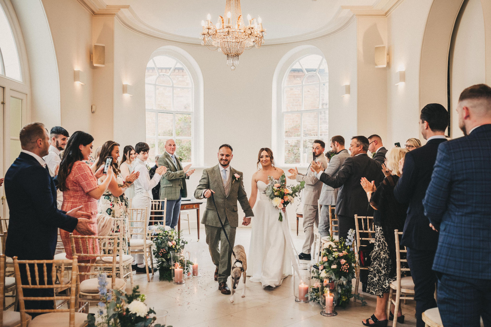 A bride and groom walk down the aisle in a white Georgian room with chandeliers with a whippet on a lead whilst their guests clap and cheer