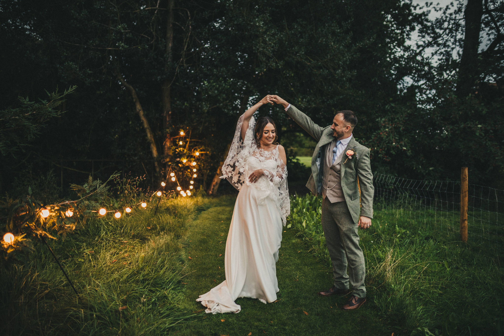A bride and groom dance on the grass under the shade of a large pine tree at dusk lit up by festoon lights looped along a mown path