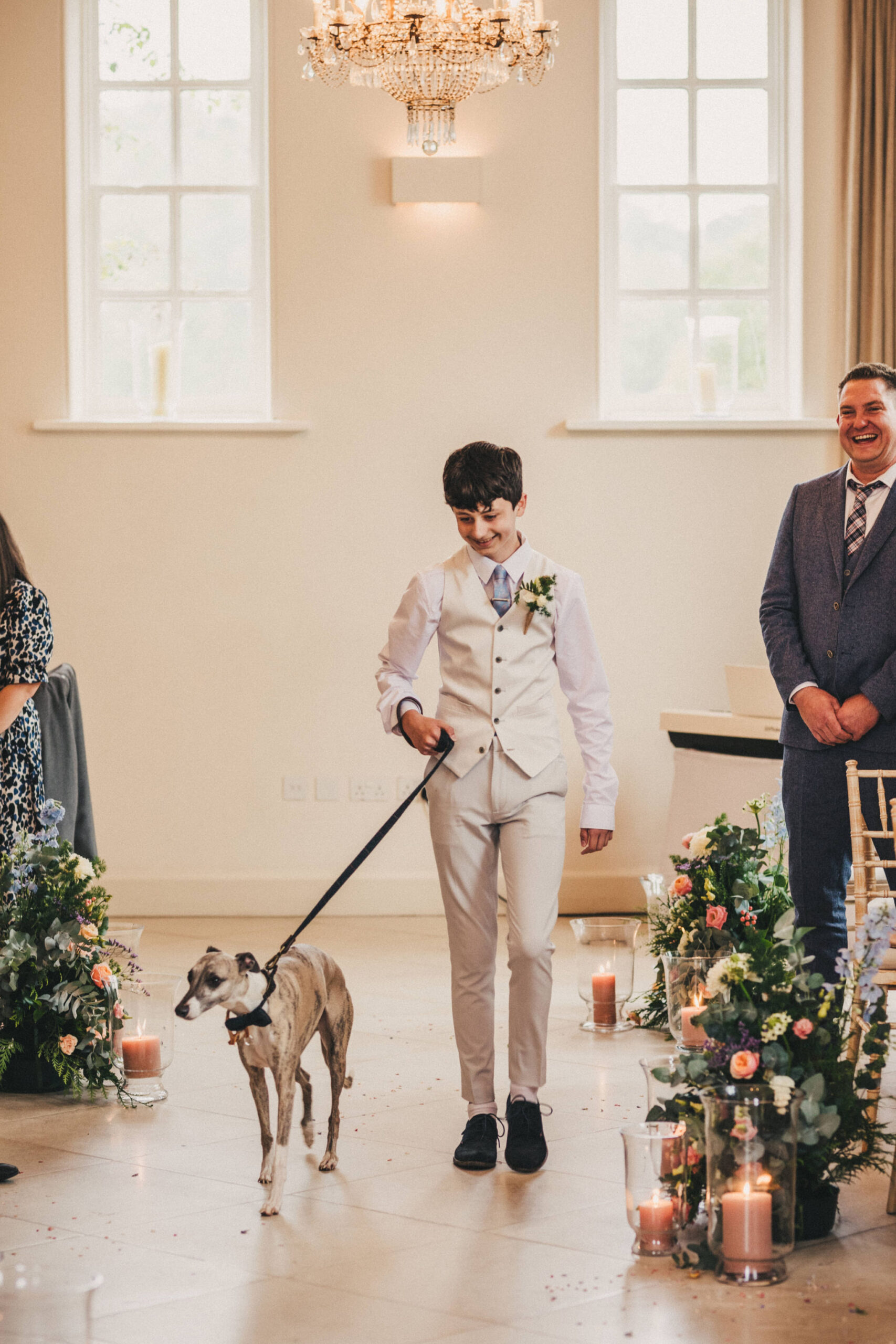 A teenage boy with a white wedding suit walks a grey whippet dog down the aisle of a room with a white flagstone floor with floral arrangement set on the floor at intervals along the rows of chairs