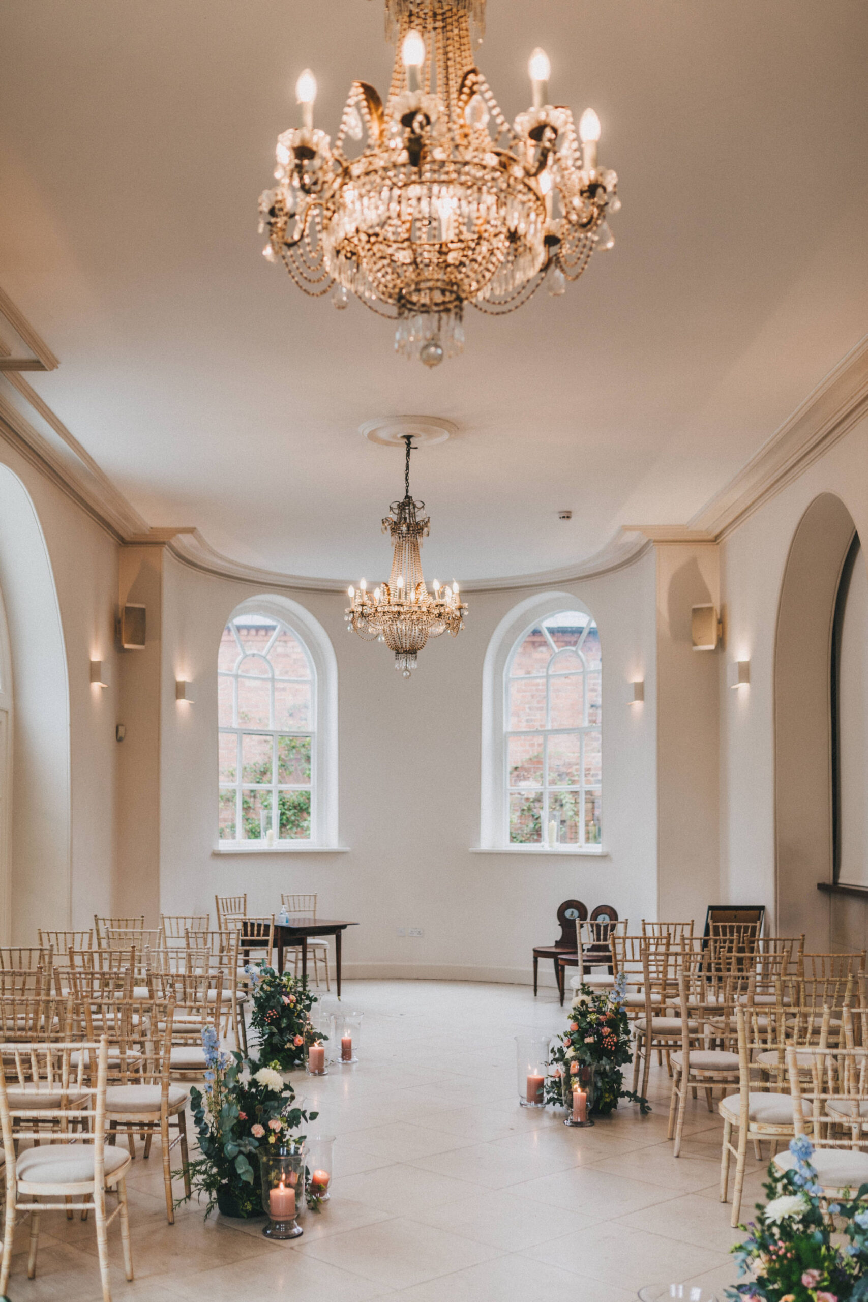 An empty wedding ceremony room with high ceilings and hanging chandeliers with a bow window at the far end