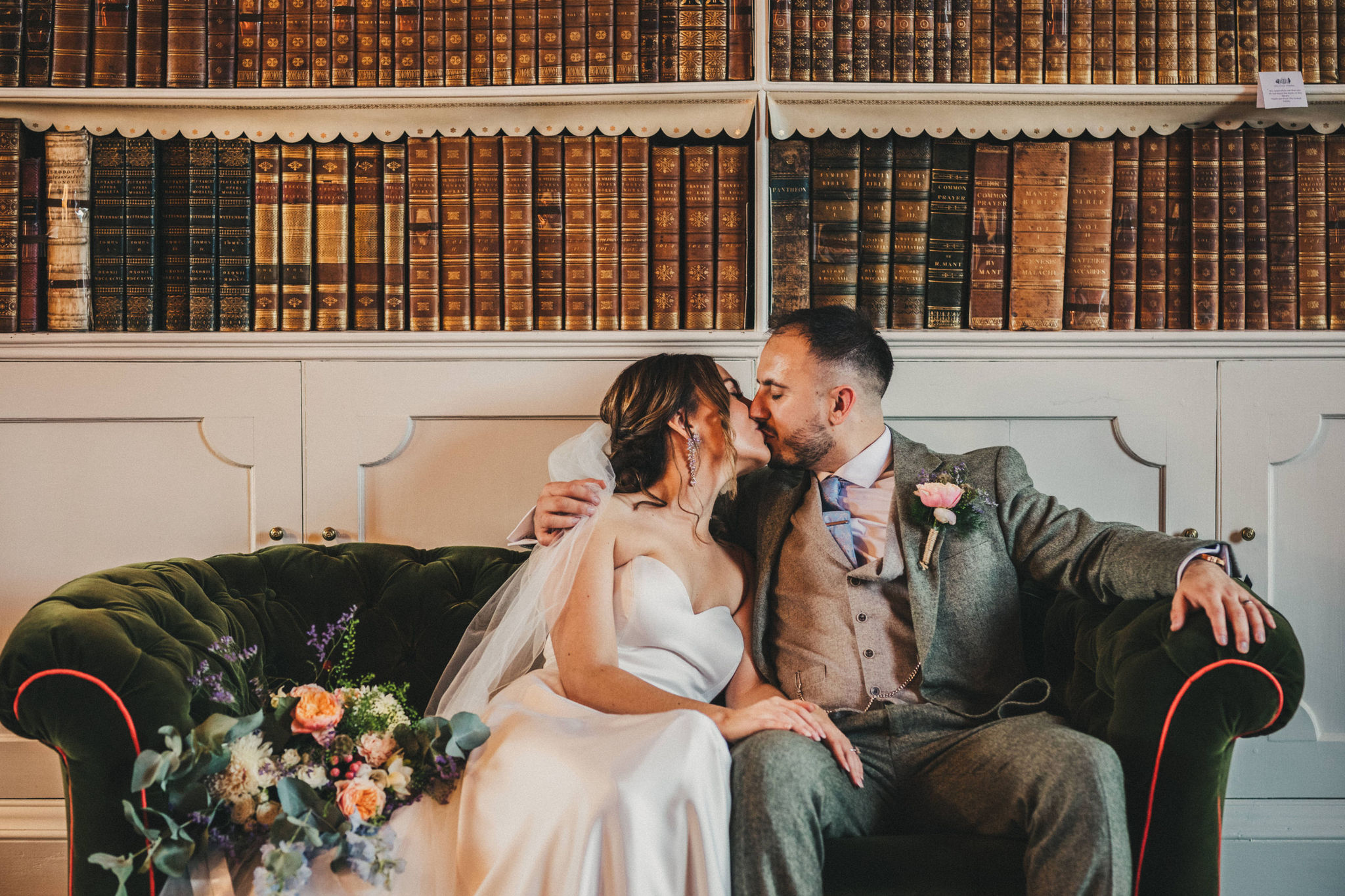 Bride and groom embracing on a dark green velvet Chesterfield sofa with bookshelves of books behind them
