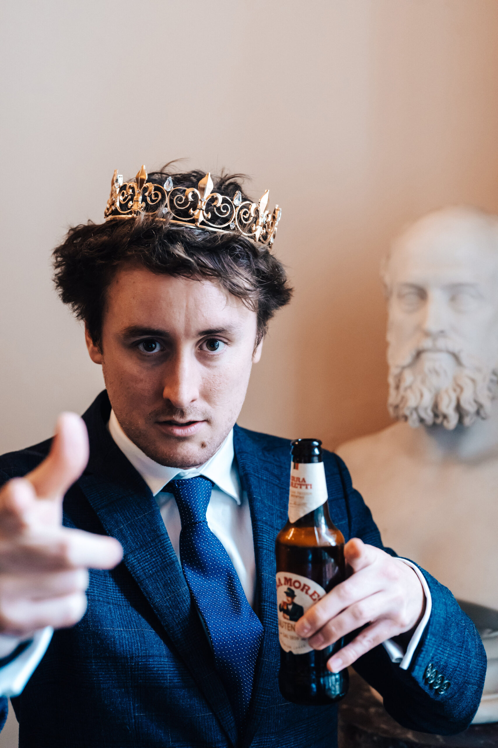 A man wearing a blue formal suit and blue tie with a bottle of beer in his hands and a gold paper crown on his head