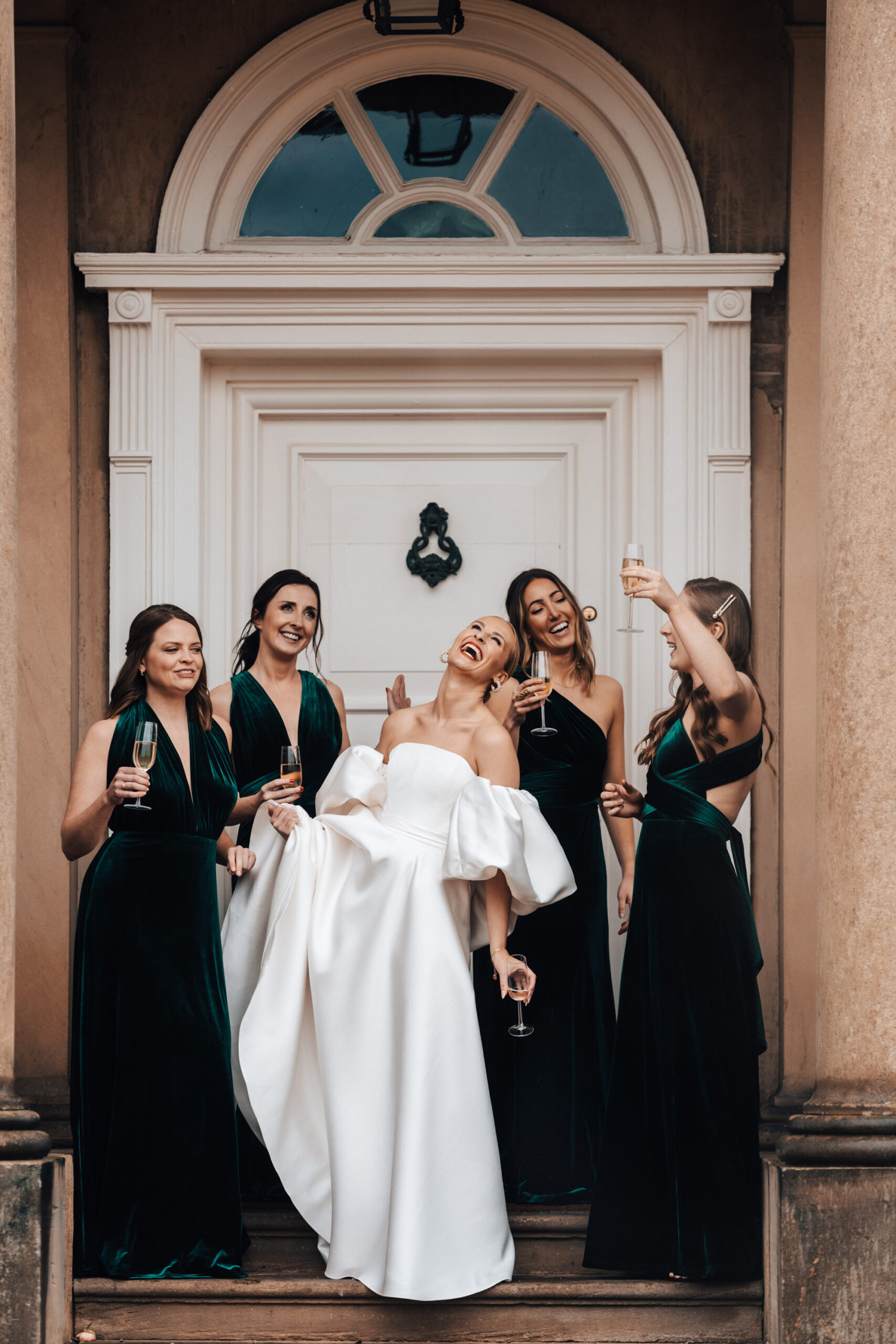 Bride and bridesmaids wearing dark green floor length dresses and modern voluminous wedding dress with large sleeves in front of a red brick Georgian Manor House