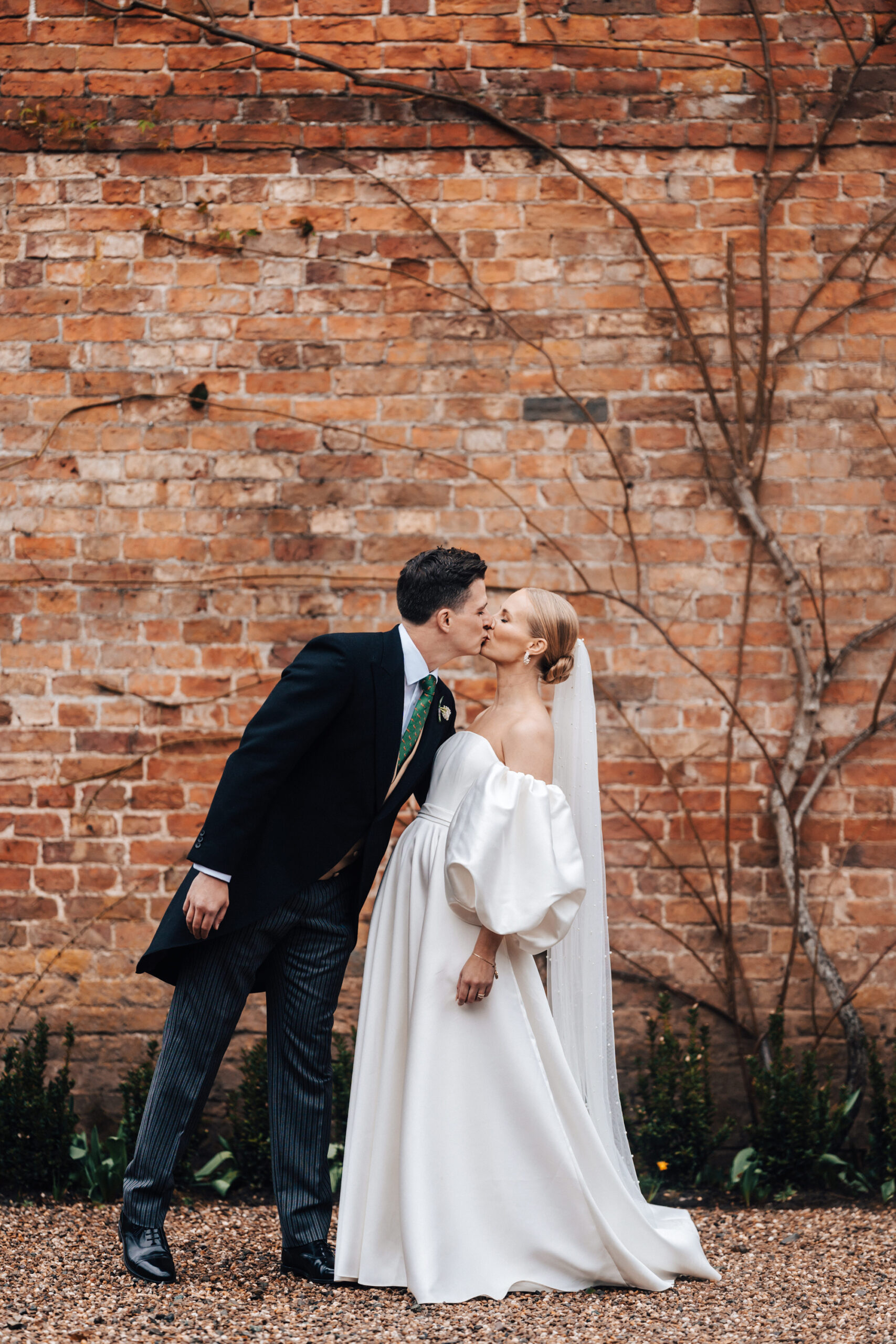 Bride and groom by red brick wall that has climbing roses growing up it to give her a kiss