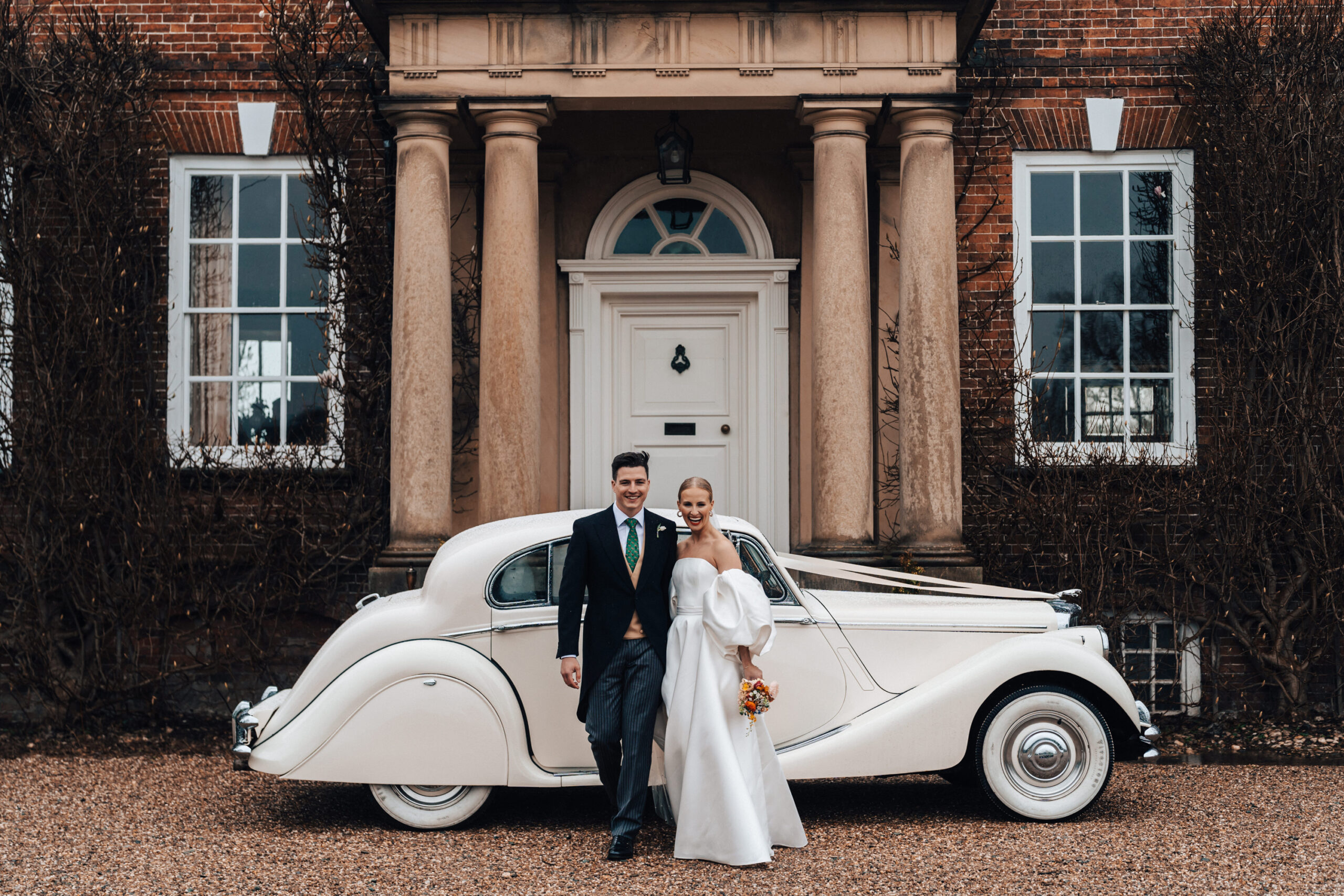 Bride and groom wearing a traditional morning suit and a modern voluminous wedding dress with large sleeves posing next to a traditional wedding car in front of a red brick Georgian Manor House