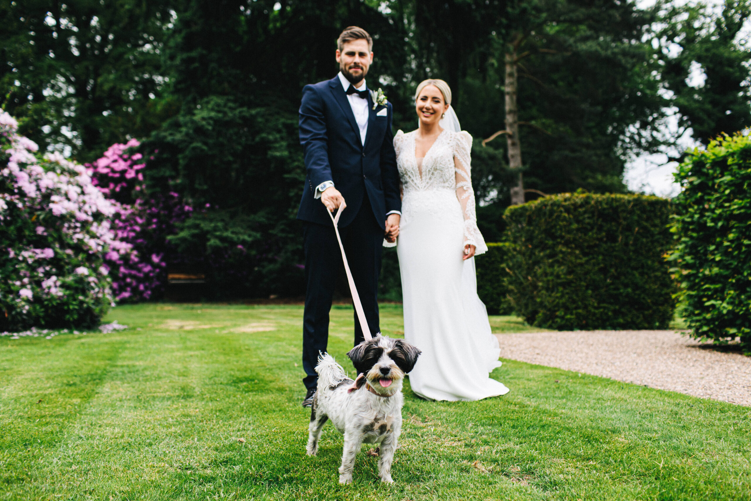 A bride in a slim fitting tulle wedding dress and a groom wearing a black tie standing in the background whilst a small black and white dog poses for the camera on a pink ribbon lead