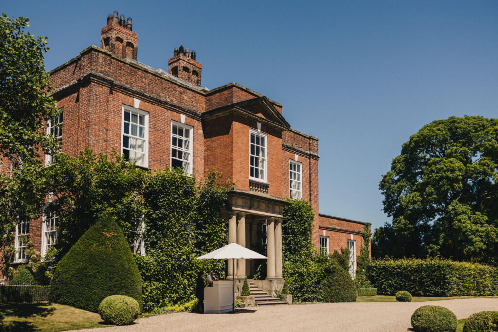 A view of a red brick Georgian Manor House flanked by large oak and beech trees set against a blazing blue Summer Sky