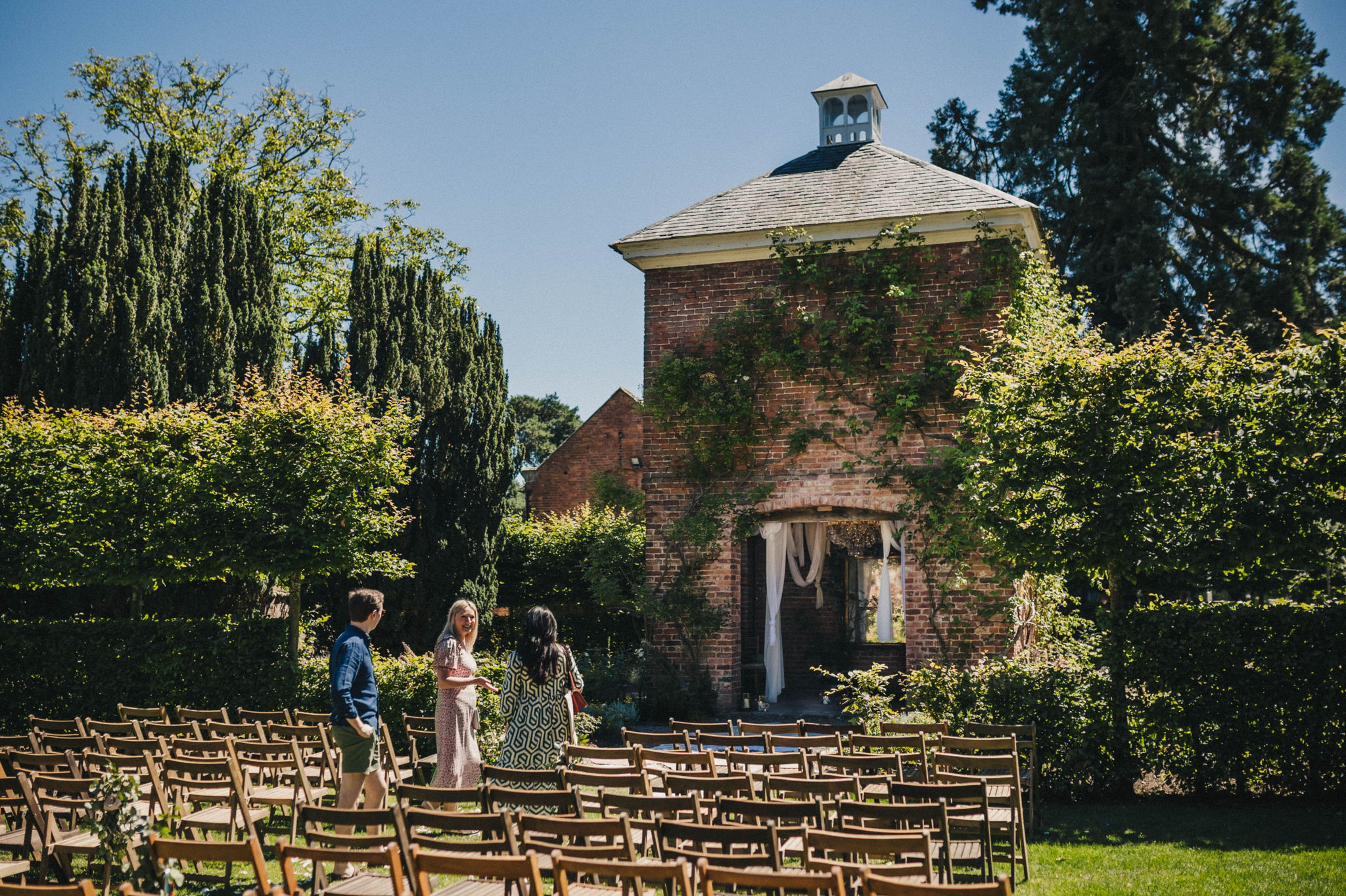 A large red brick dovecote sits amongst landscaped gardens under a clear blue sky with climbing pink roses up the height of its walls