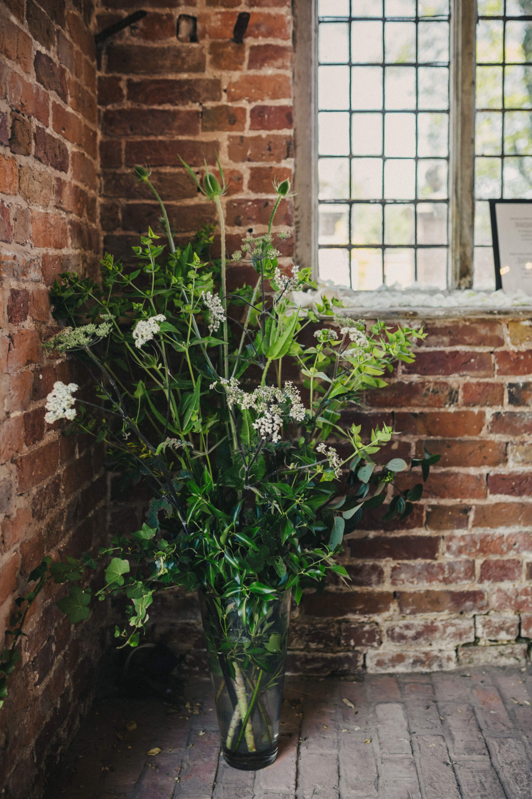 A lush green floral arrangement with white accents sits on the floor of a building that has exposed red brick walls and a wooden untreated floor