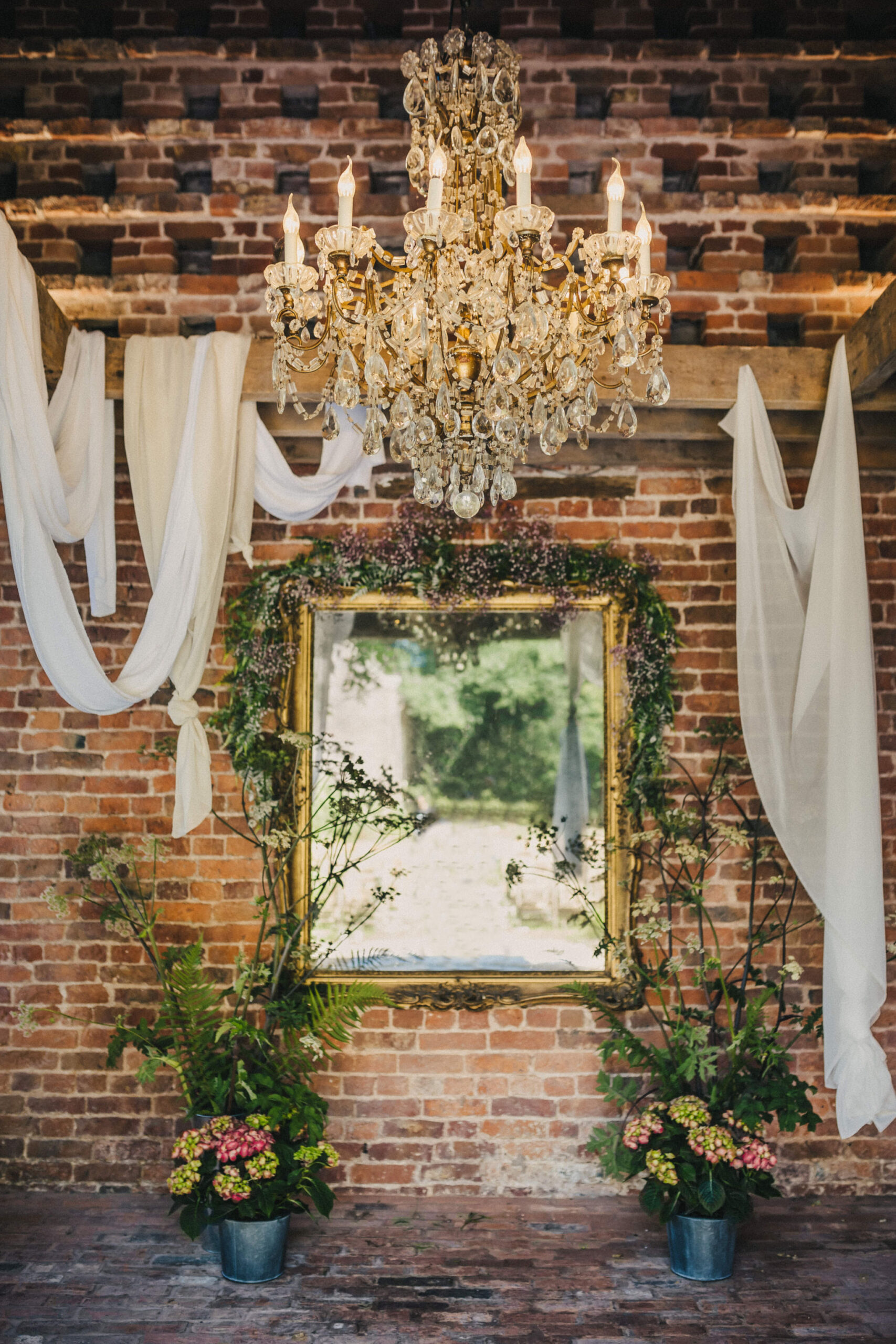 An image of a gold ornate mirror hanging on an exposed red brick wall which is draped in green foliage with a crystal chandelier hanging above