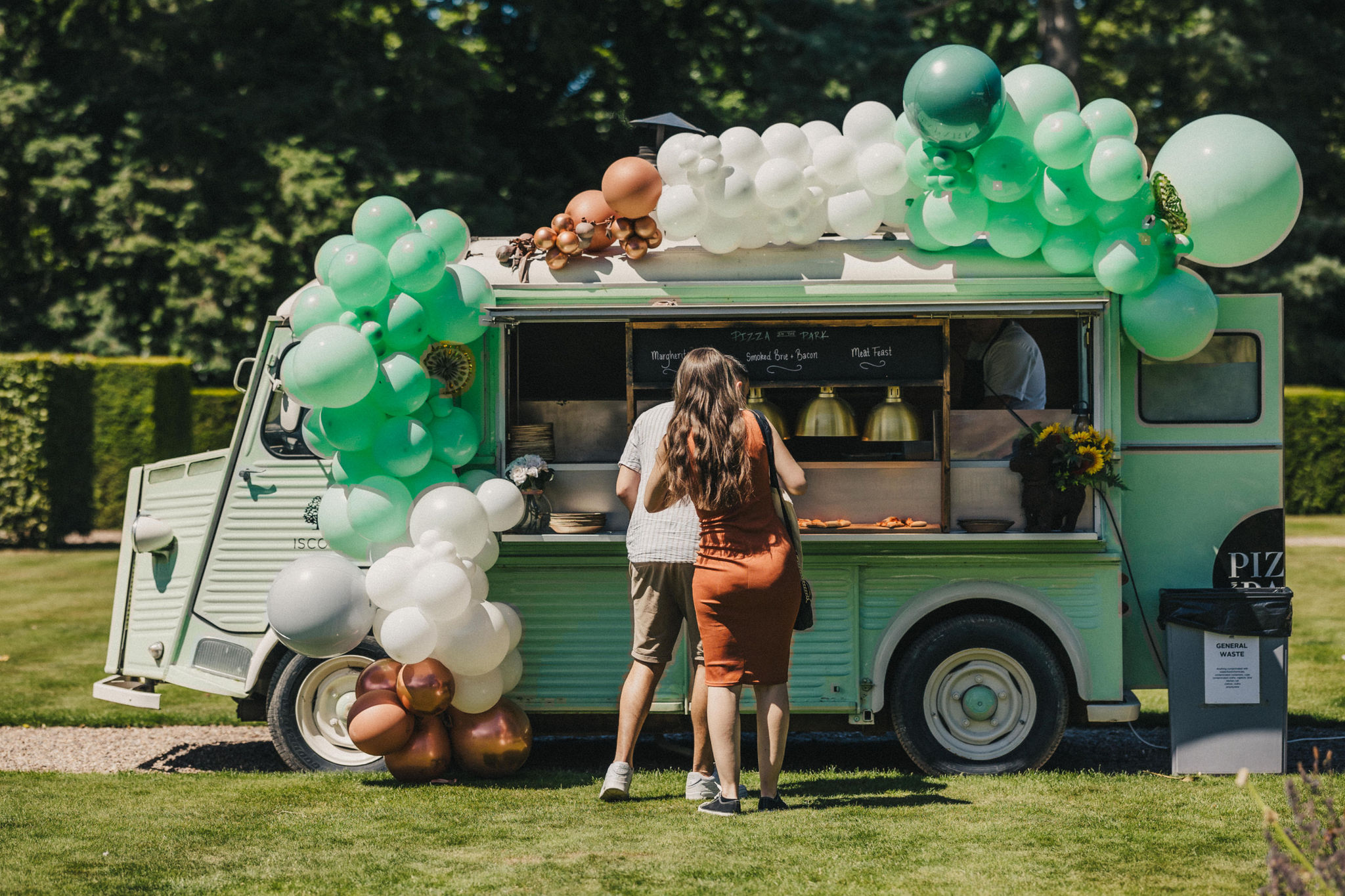 A mint green vintage food truck serves two people freshly made pizza under blue skies