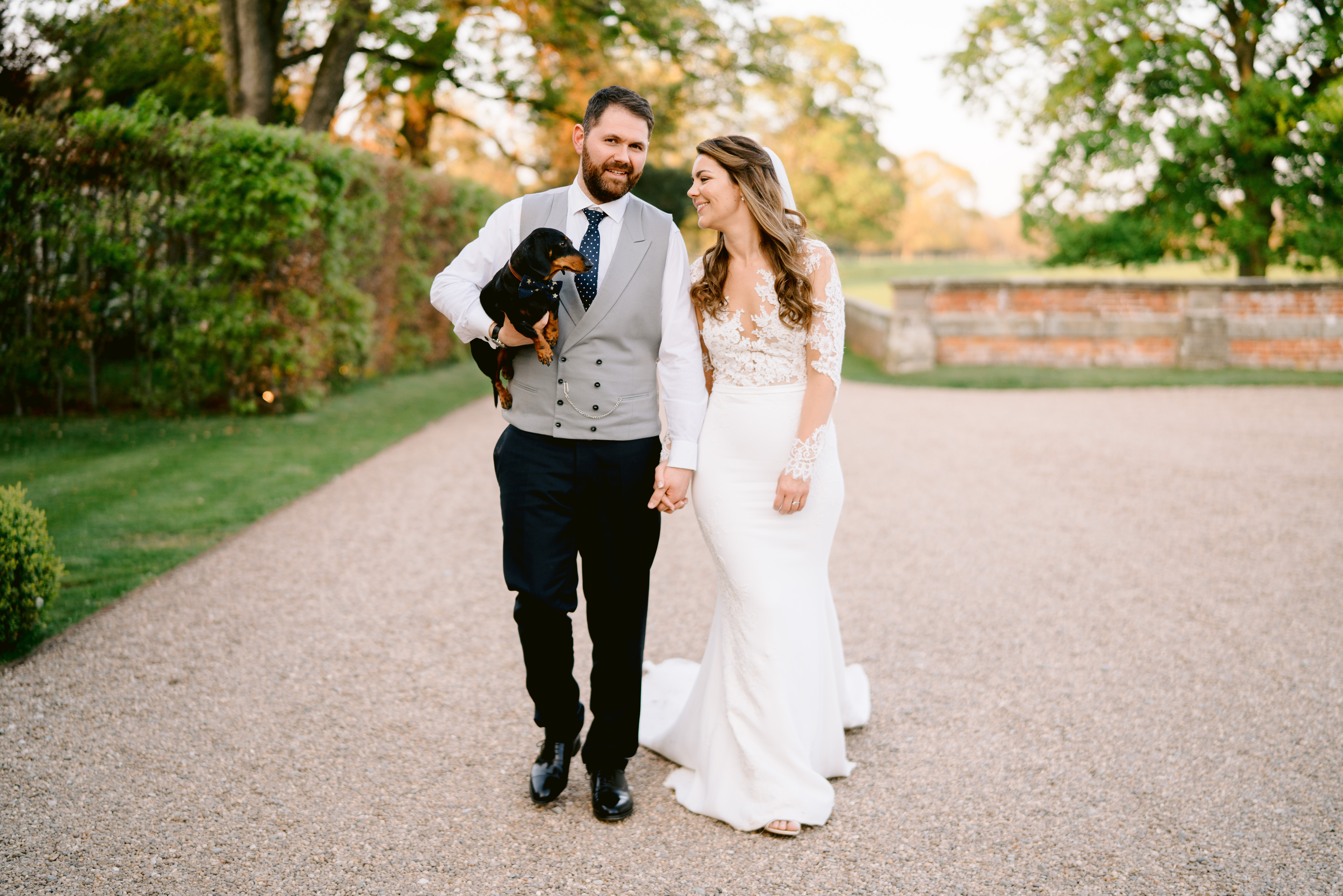 A bride holds hands with her groom who is wearing a grey waistcoat and trousers holding a Black and Tan sausage dog