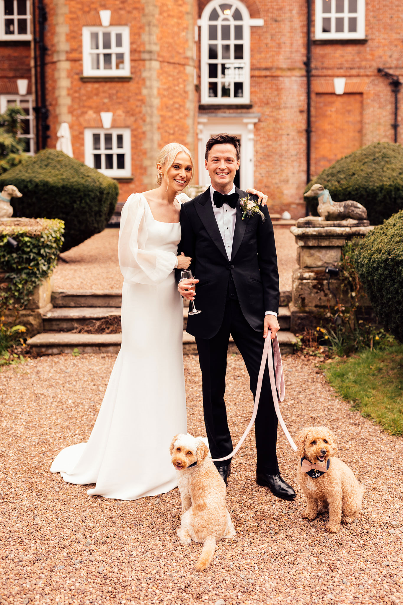 A bride in a slim fitting tulle wedding dress and a groom wearing a black tie walk in landscaped gardens with two pet terrier dogs on ribboned leads