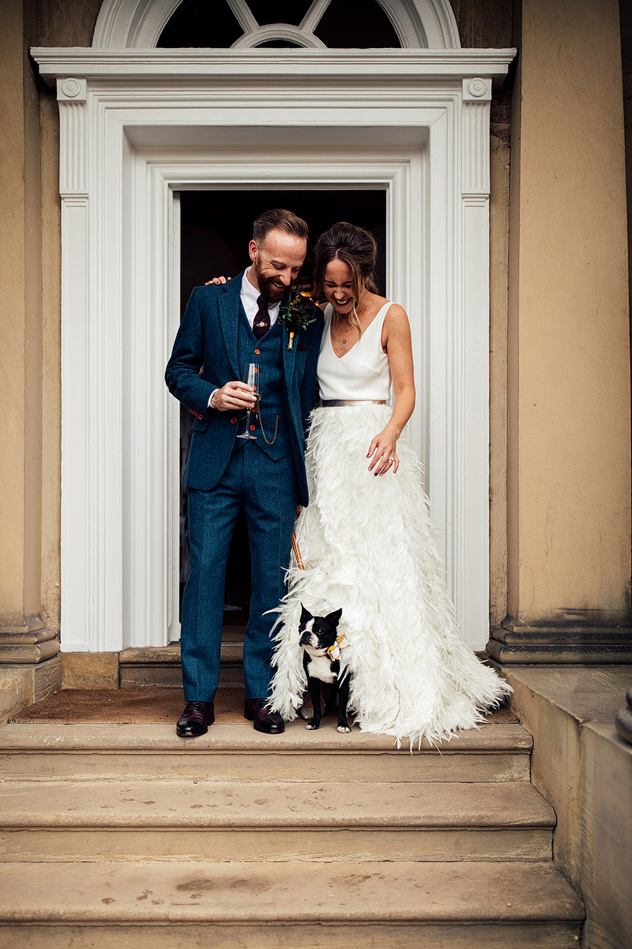 A bride in a feathered wedding dress and a groom wearing a blue suit stand at the top of some York Stone steps by an open doorway with their French Bulldog at their feet