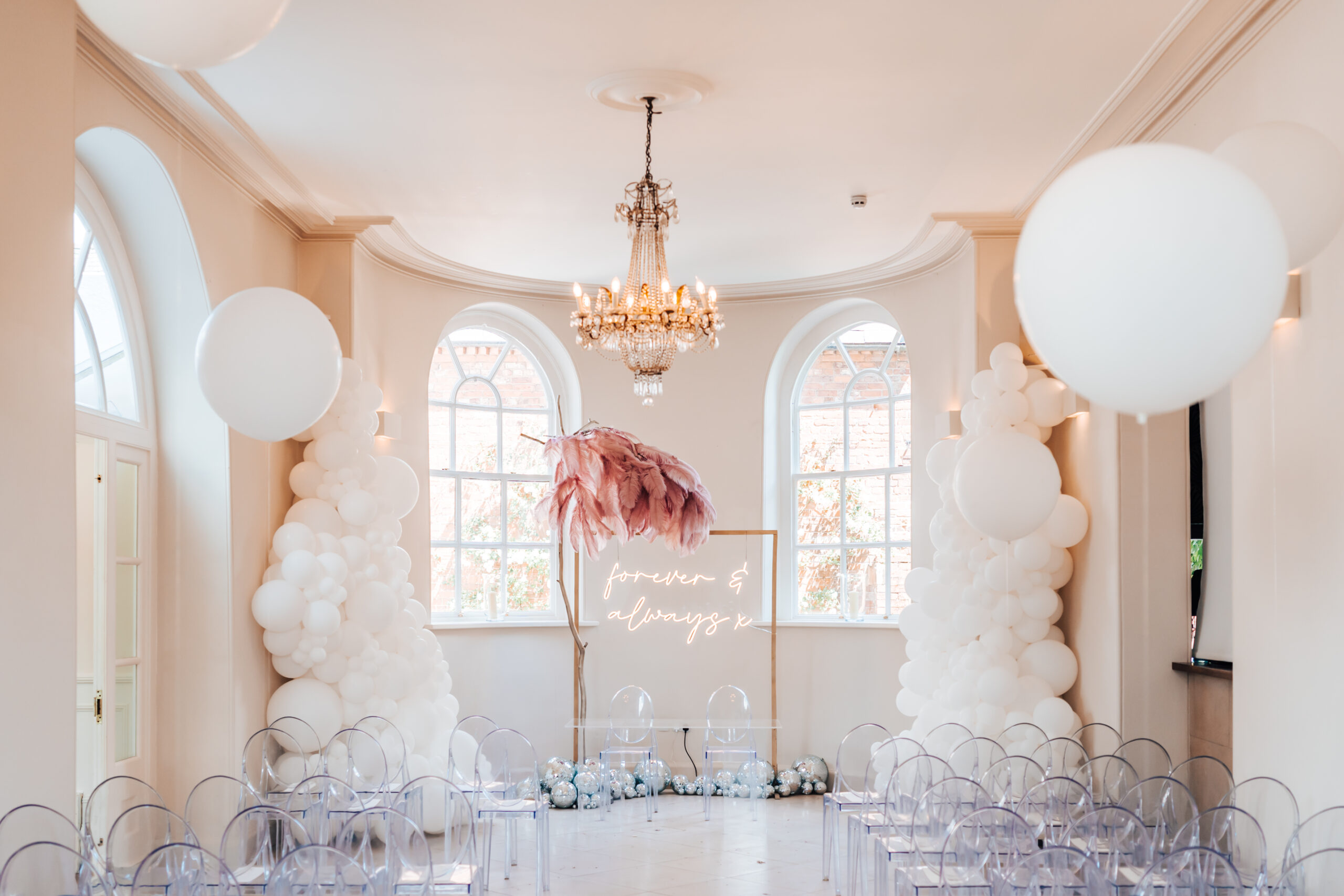 A wedding aisle altarpiece consisting of a neon sign saying forever and always flanked by an ostrich feather tree and hundreds of silver toned mirrorball and supersized white balloons