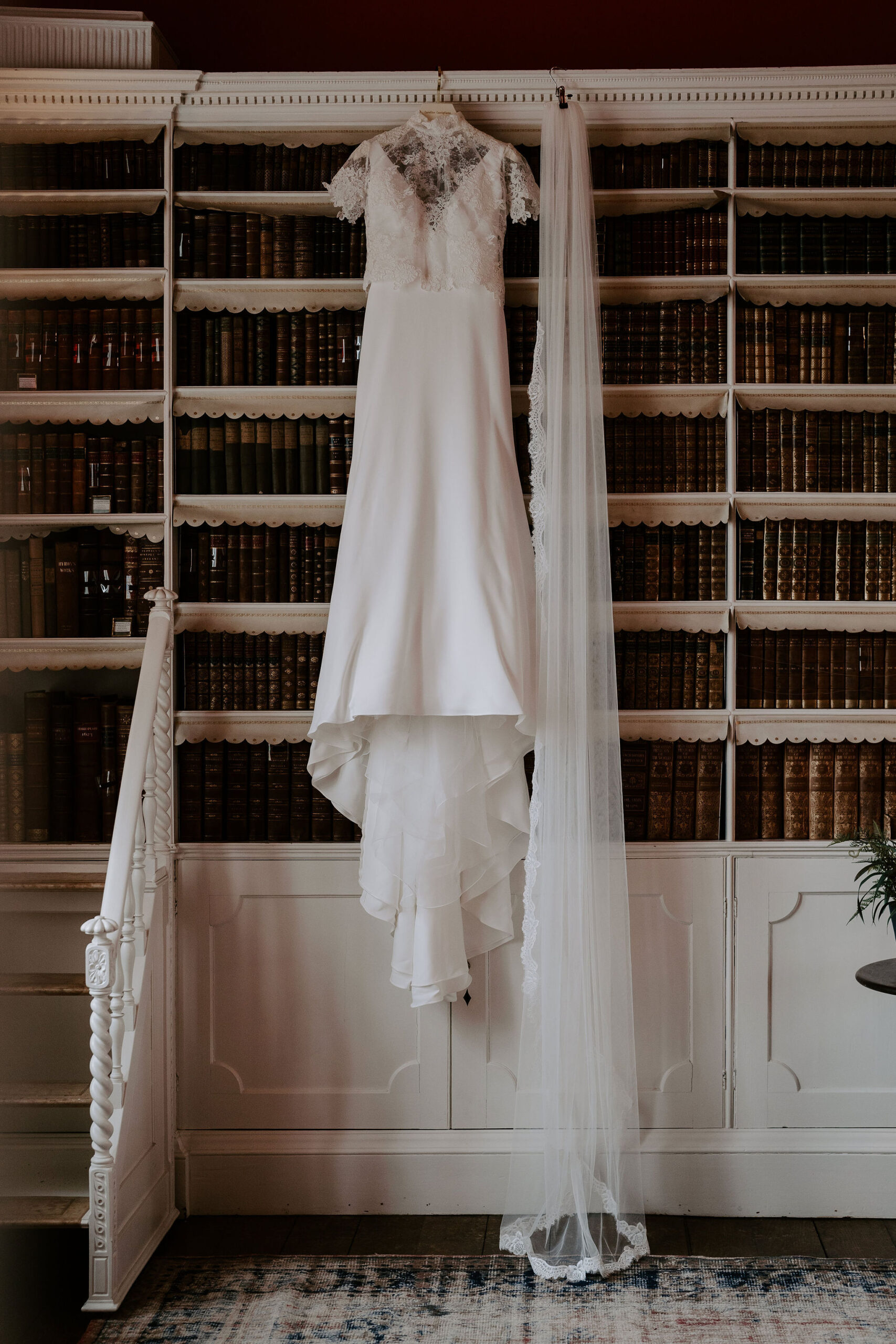 A white lace wedding gown hangs next to a long veil on a set of antique white bookshelves that have antique leather bound books upon them