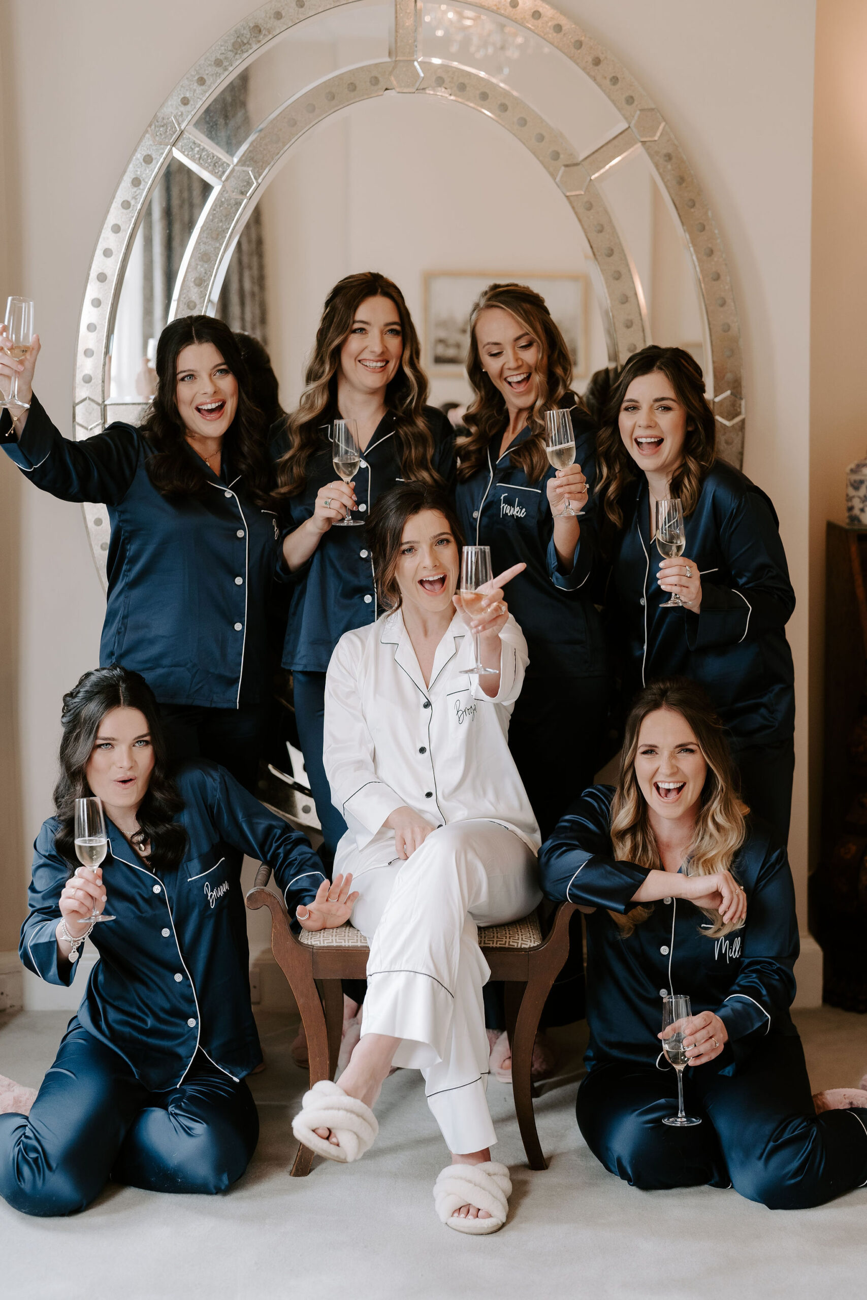 Six bridesmaids wearing navy silk pyjamas with white trimmings surround a bride wearing a white silk pair of pyjamas holding champagne glasses and laughing at the camera