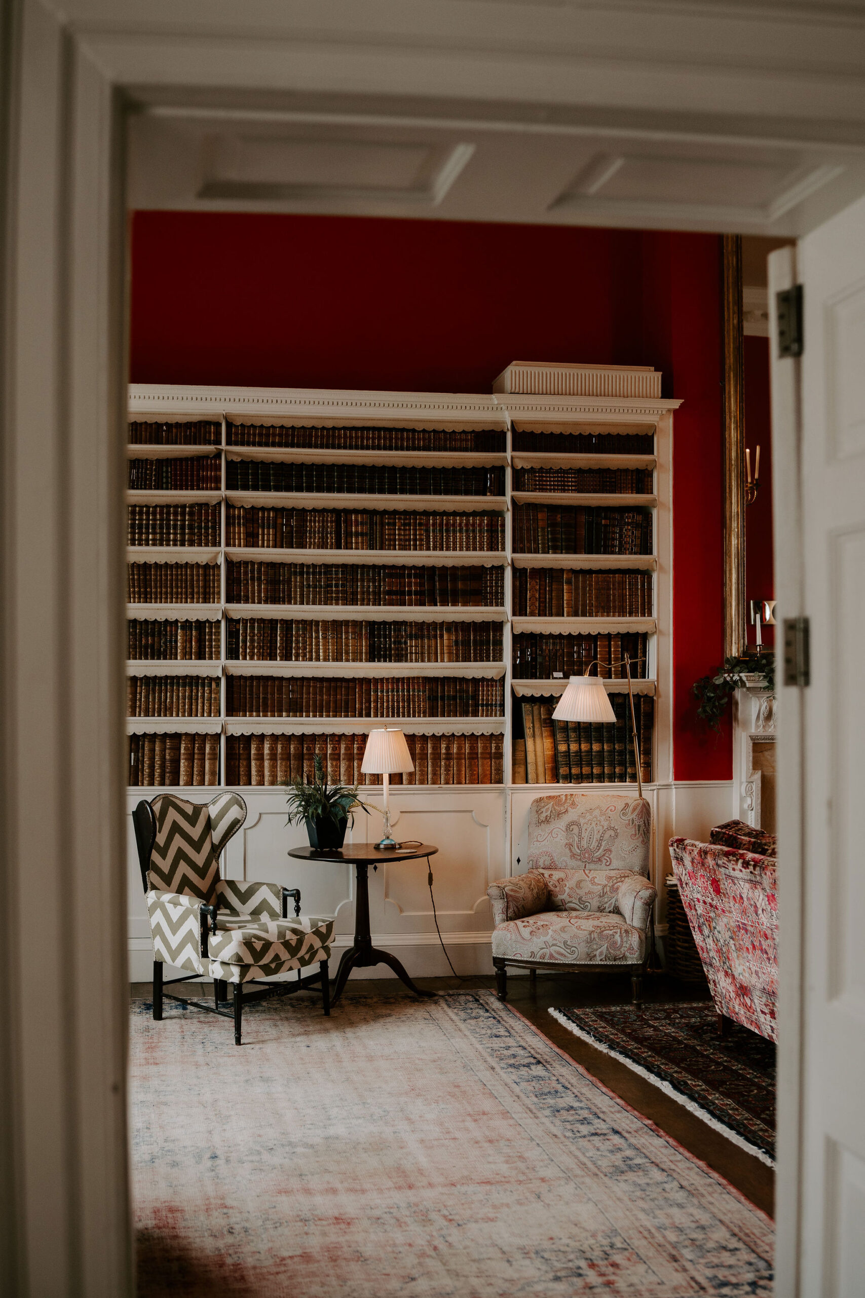 A glimpse into an old library with antique books lined up on white bookshelves. Two armchairs covered in paisley and checks are positioned in front of the bookshelves with a small wooden side table between them on which is a lamp and a plant