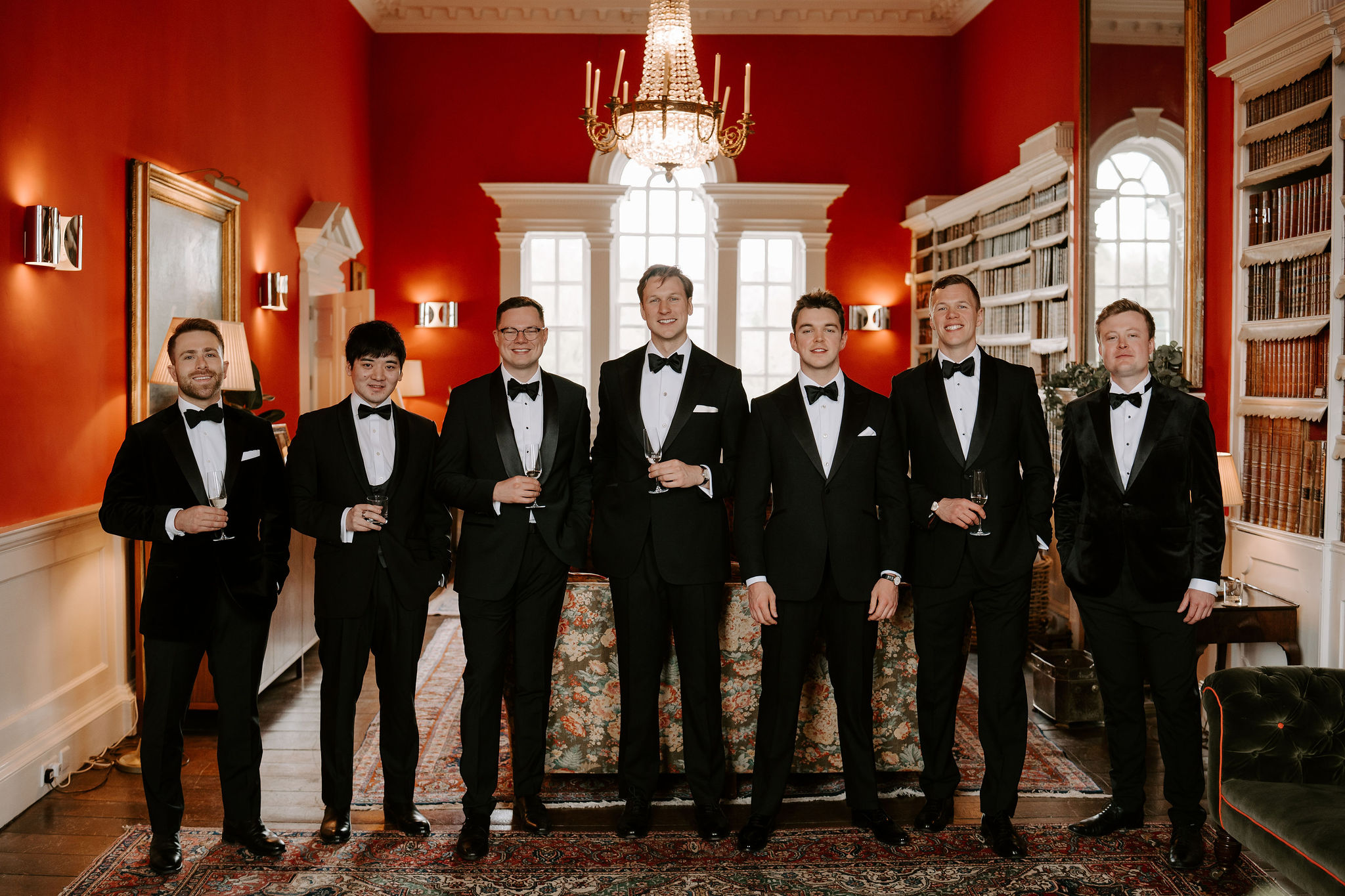 Seven men in black tie line up in a row holding glasses of champagne in an antique library painted red