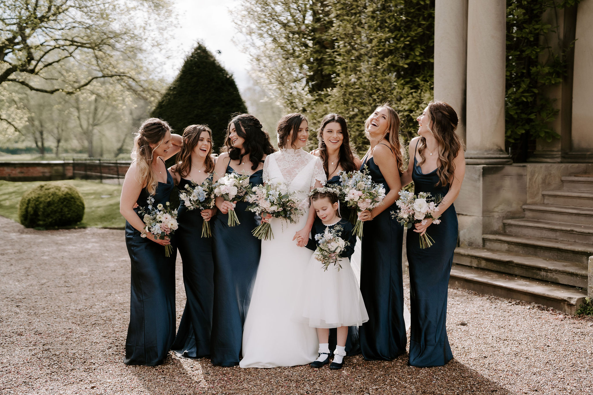Six bridesmaids wearing navy silk slip dresses surround a bride wearing a white lace short sleeved dress holding pastel bouquets
