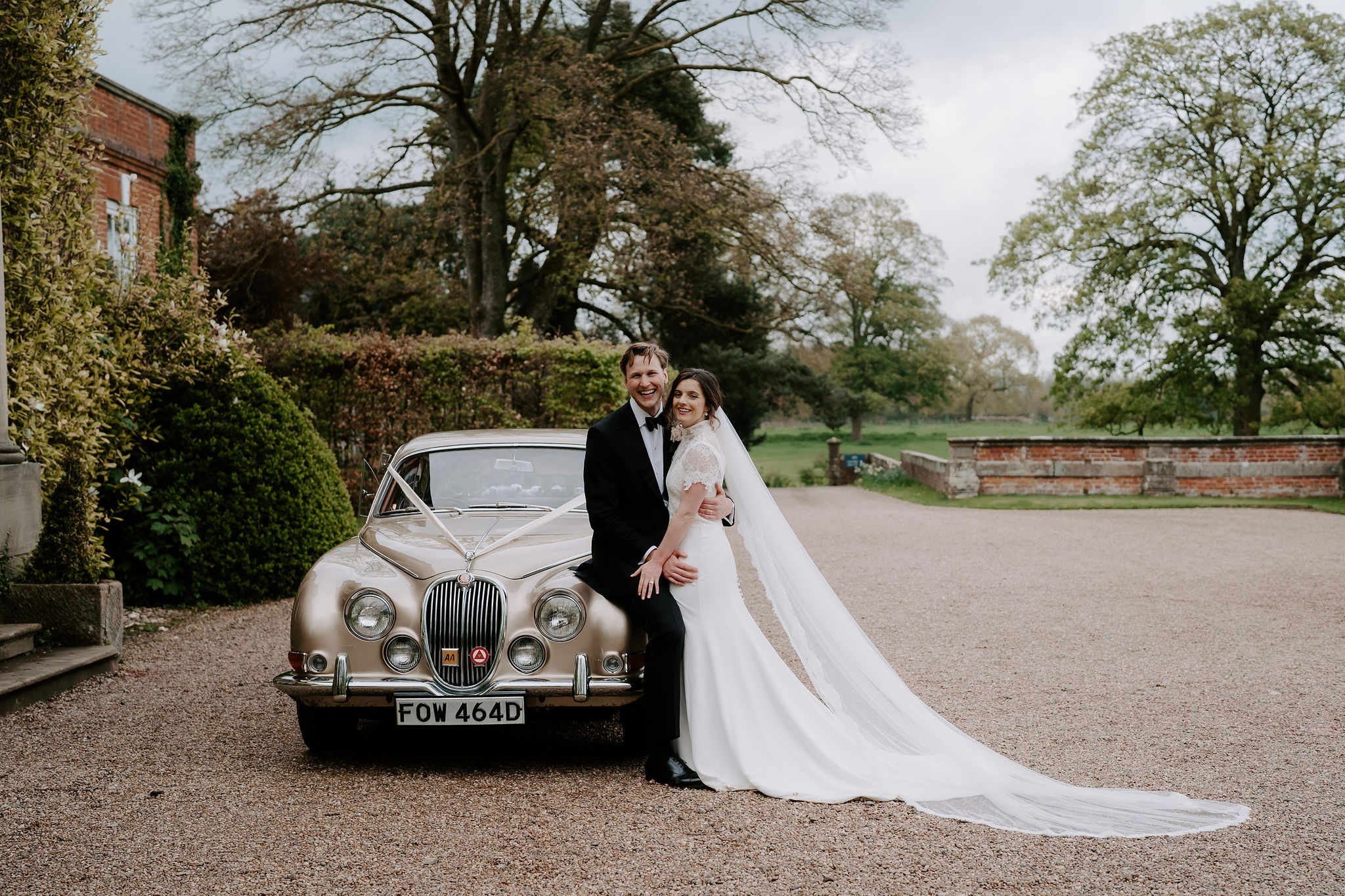 A groom wearing black tie and a bride wearing a lace short sleeved dress pose next to an old vintage car which is parked outside a redbrick Georgian mansion