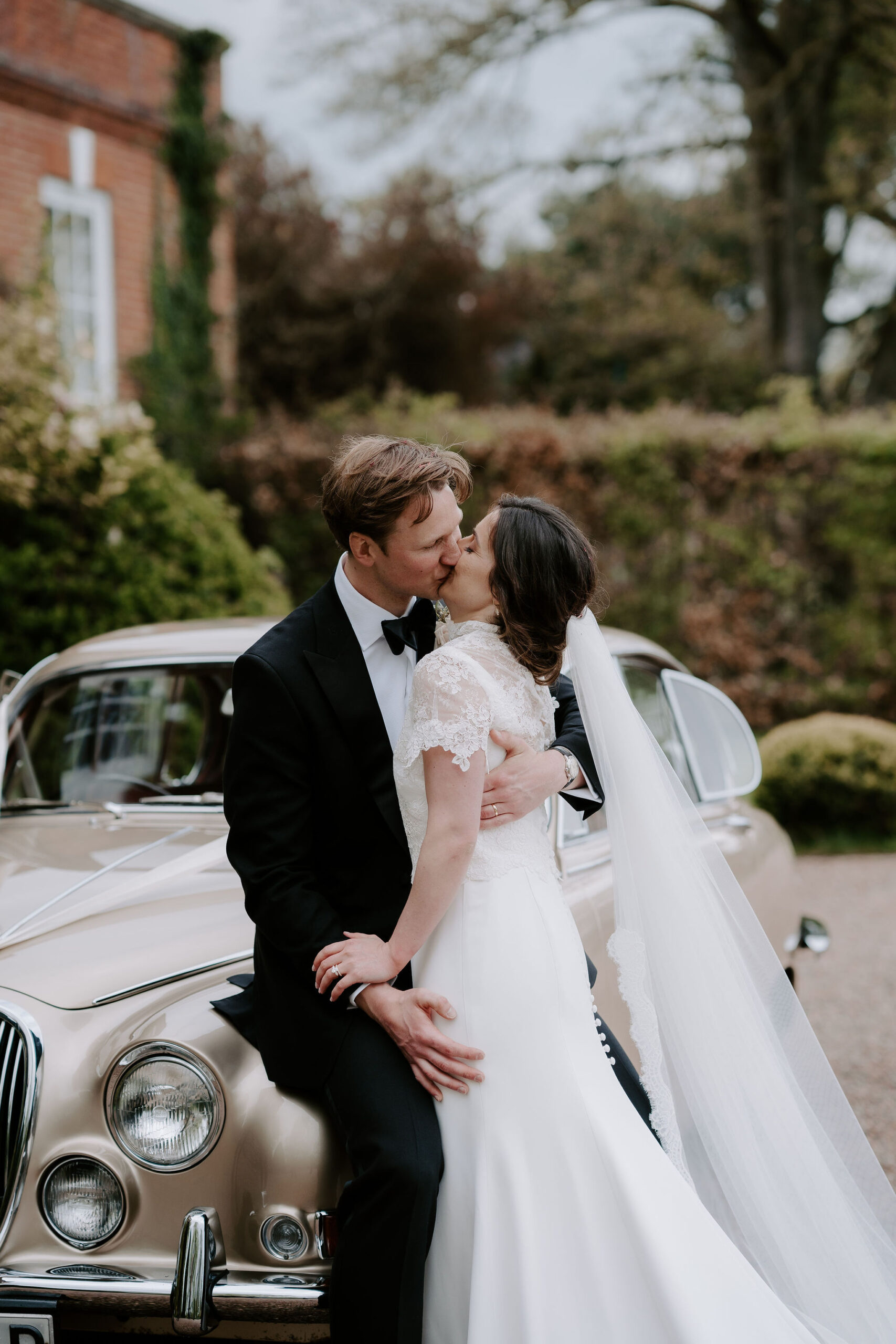 A groom wearing black tie and a bride wearing a lace short sleeved dress kiss next to an old vintage car