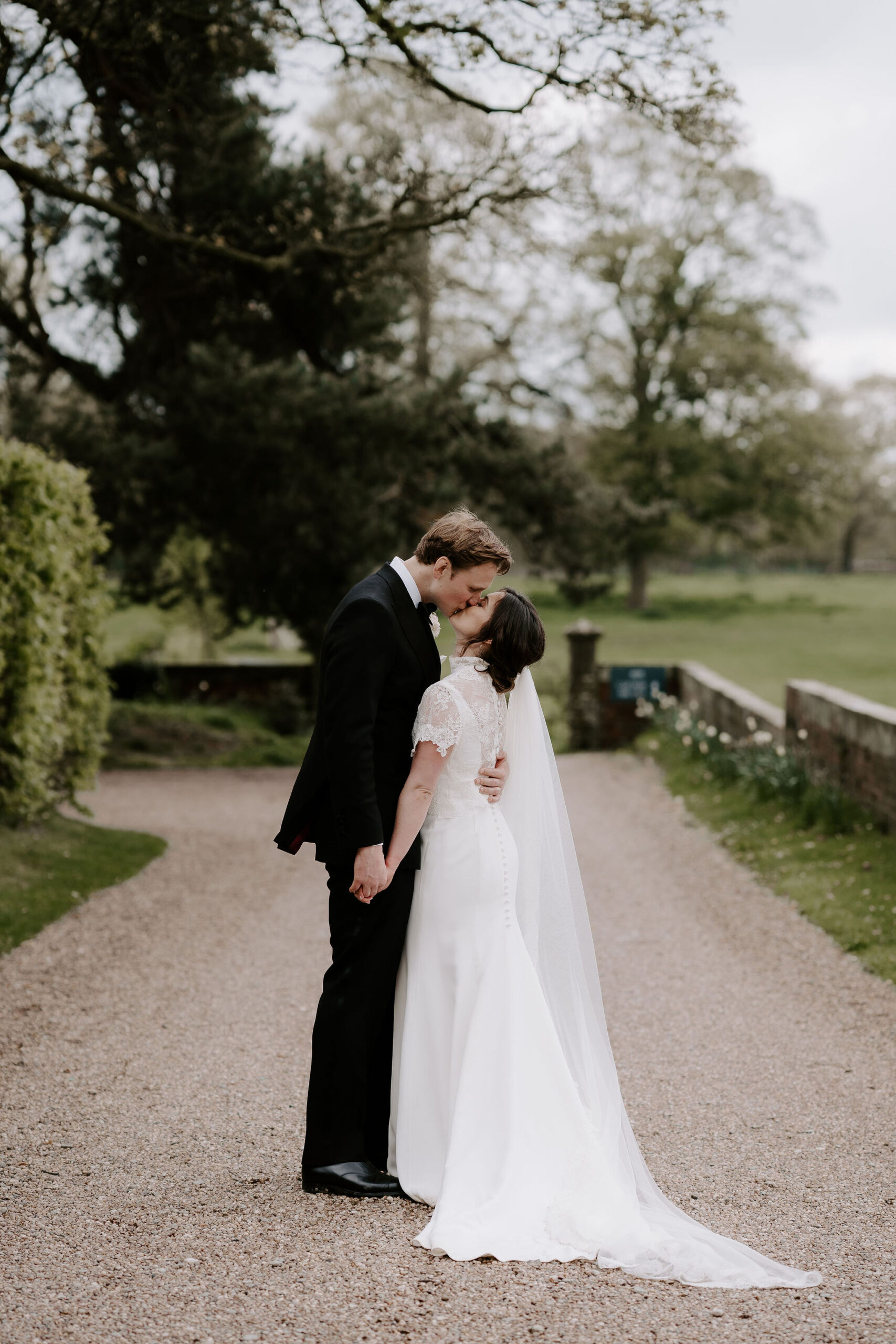 A bride and groom wearing black tie and a lacy dress kiss whilst standing outside on a gravel driveway