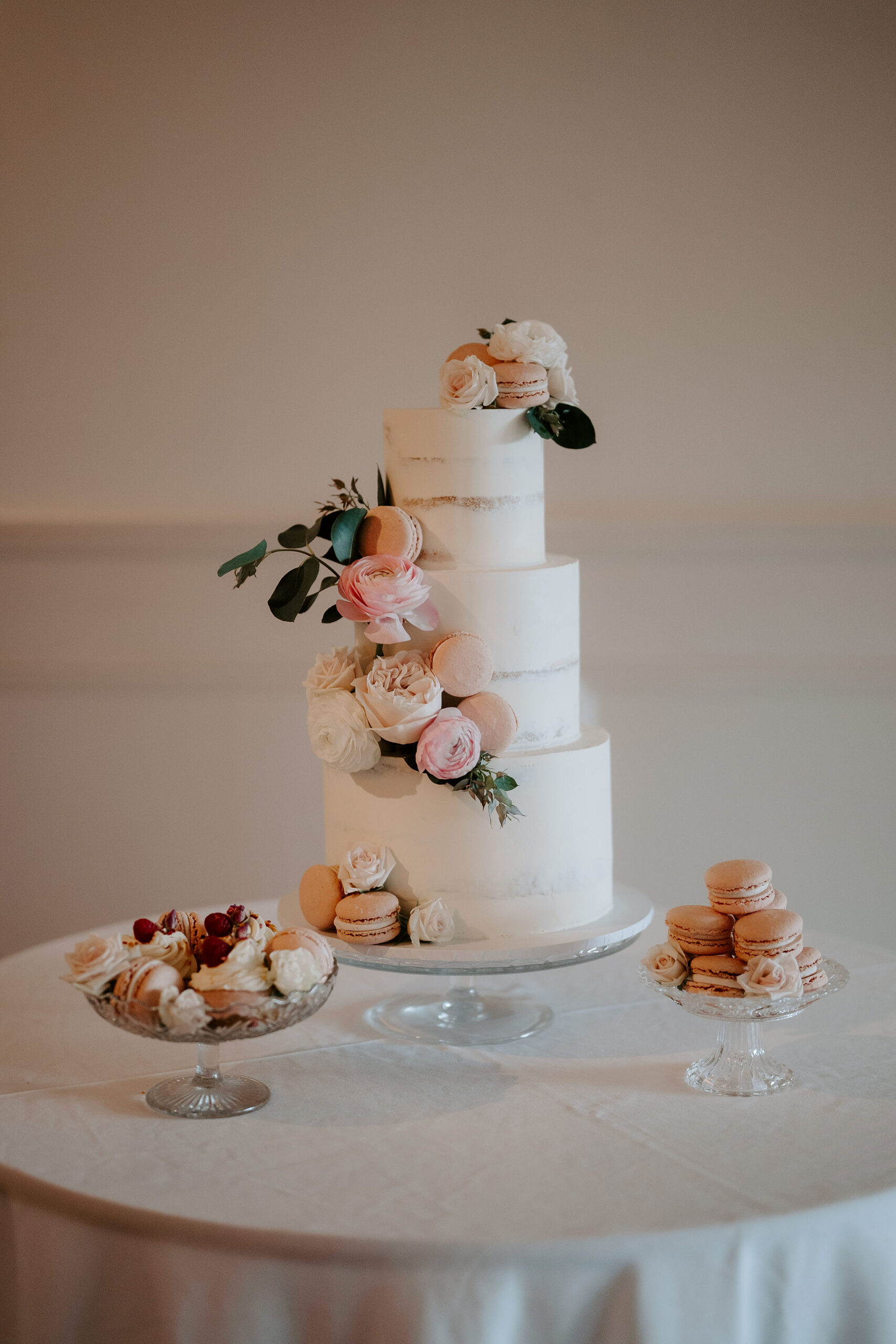 A three tiered white wedding cake festooned in roses and macarons sits on a white tablecloth with small bon bon dishes pilled with macarons