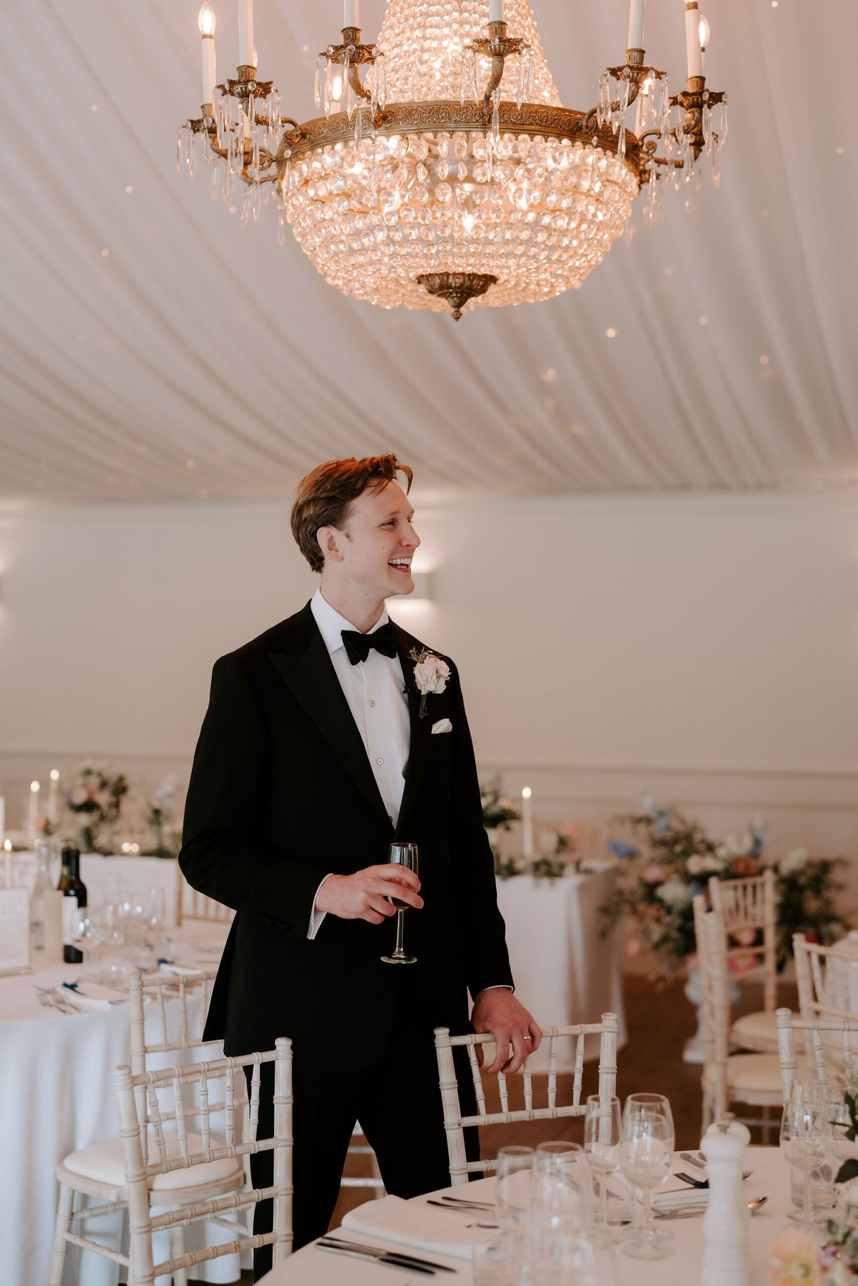 A groom in black tie stands underneath a chandelier in a marquee that has been set up for a wedding reception