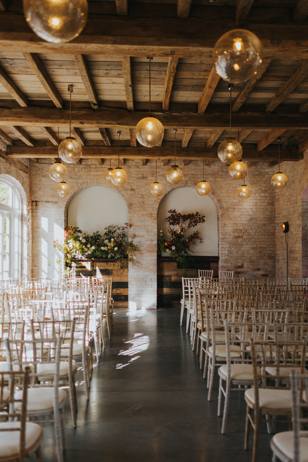 Rows of distressed white wooden chairs are lined up with spacing for an aisle to run between them inside a rustic converted coach house with exposed brick walls snd globe lights hanging from the ceiling