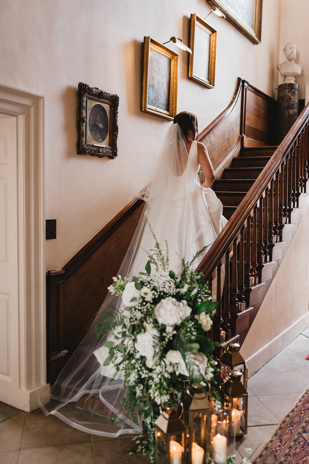 A bride in a white wedding dress and long veil walks up an antique wooden staircase that has candles arranged at the bottom and white and green flowers entwined around the bannister