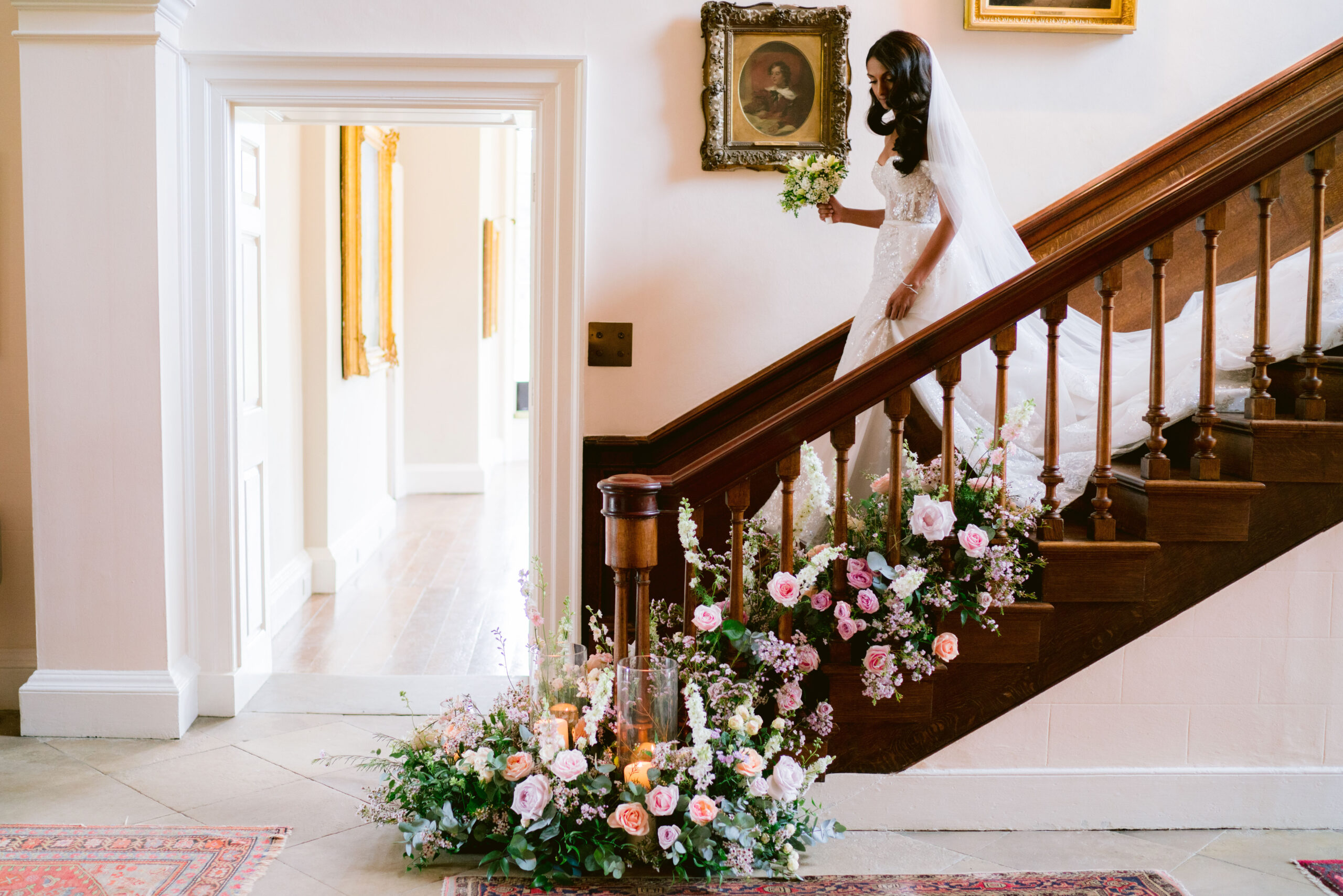 A bride descends an antique wooden staircase that has been garlanded in pastel toned spring flowers