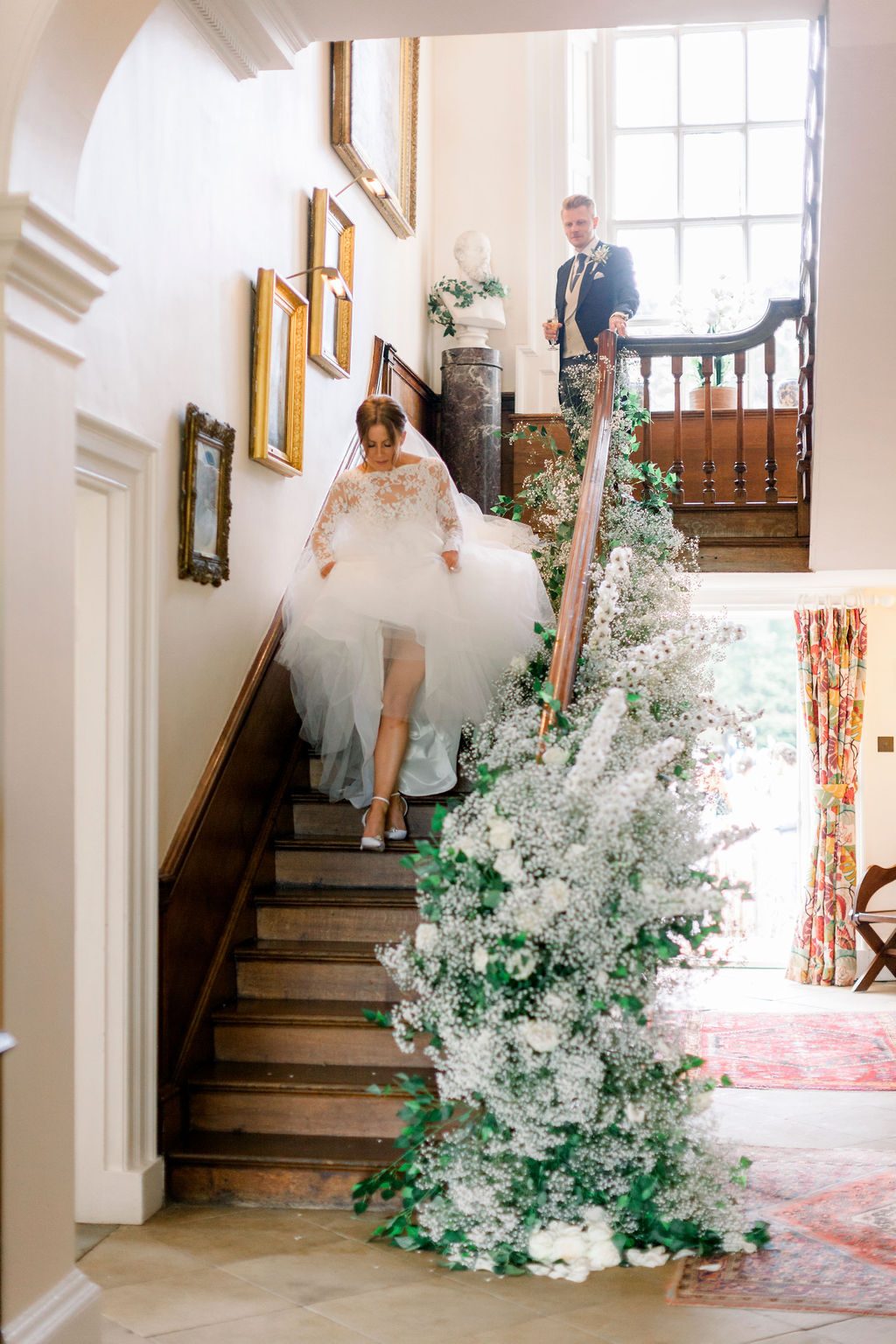 A bride in a white wedding dress and long veil walks down an antique wooden staircase that has candles arranged at the bottom and white and green flowers entwined around the bannister
