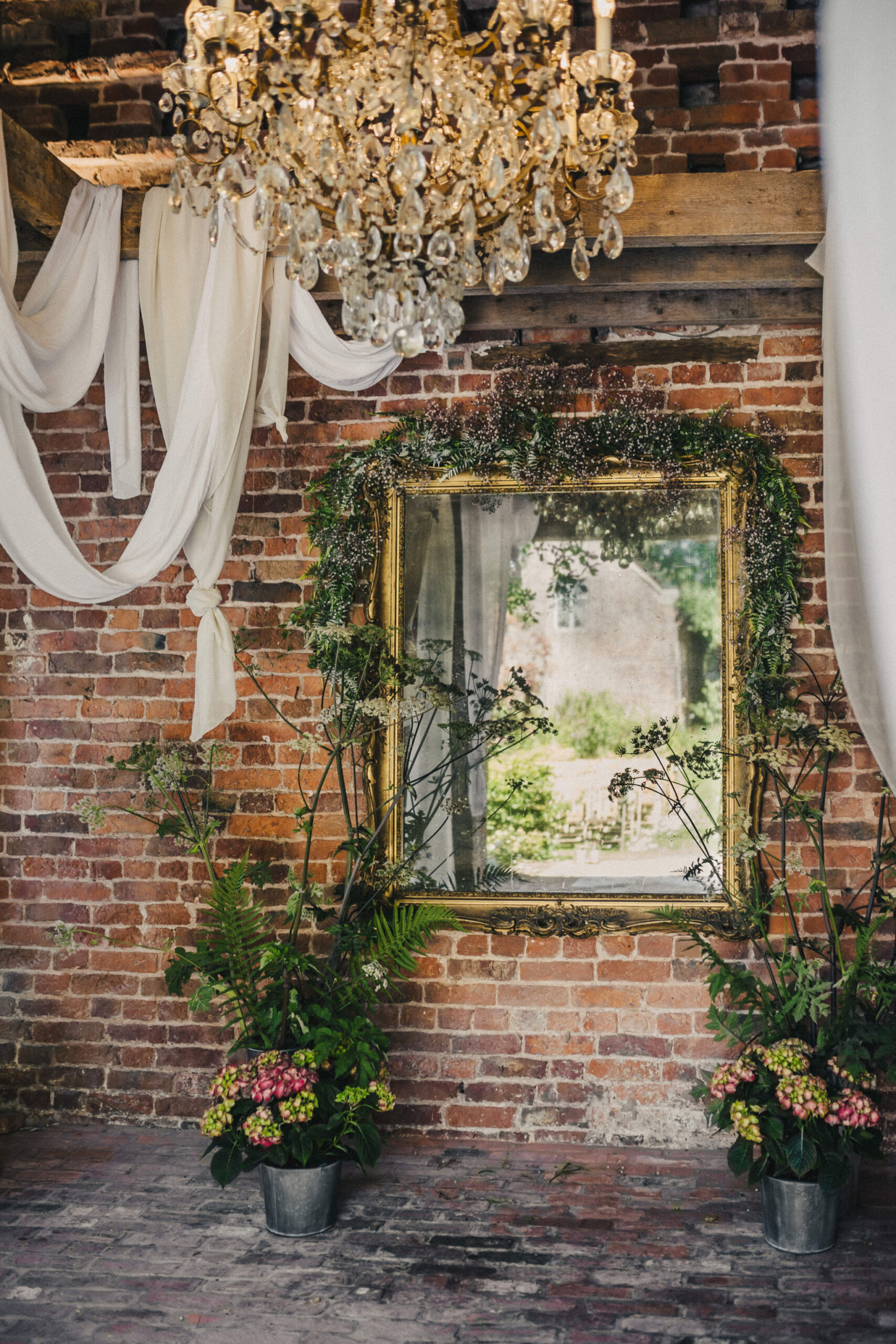 An ornate gold mirror hangs on an exposed brick wall and is surrounded by two floral displays placed on either side of the mirror on the floor