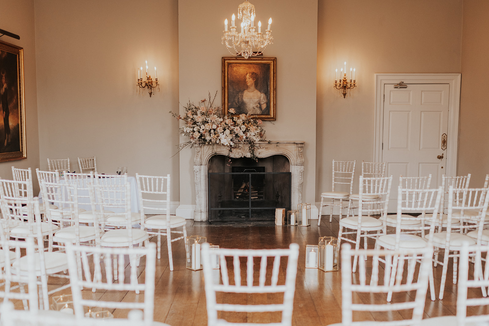 Rows of white wooden chairs are set up for a wedding ceremony in front of an elaborate stone fireplace that has an antique oil portrait of a woman hanging above it.