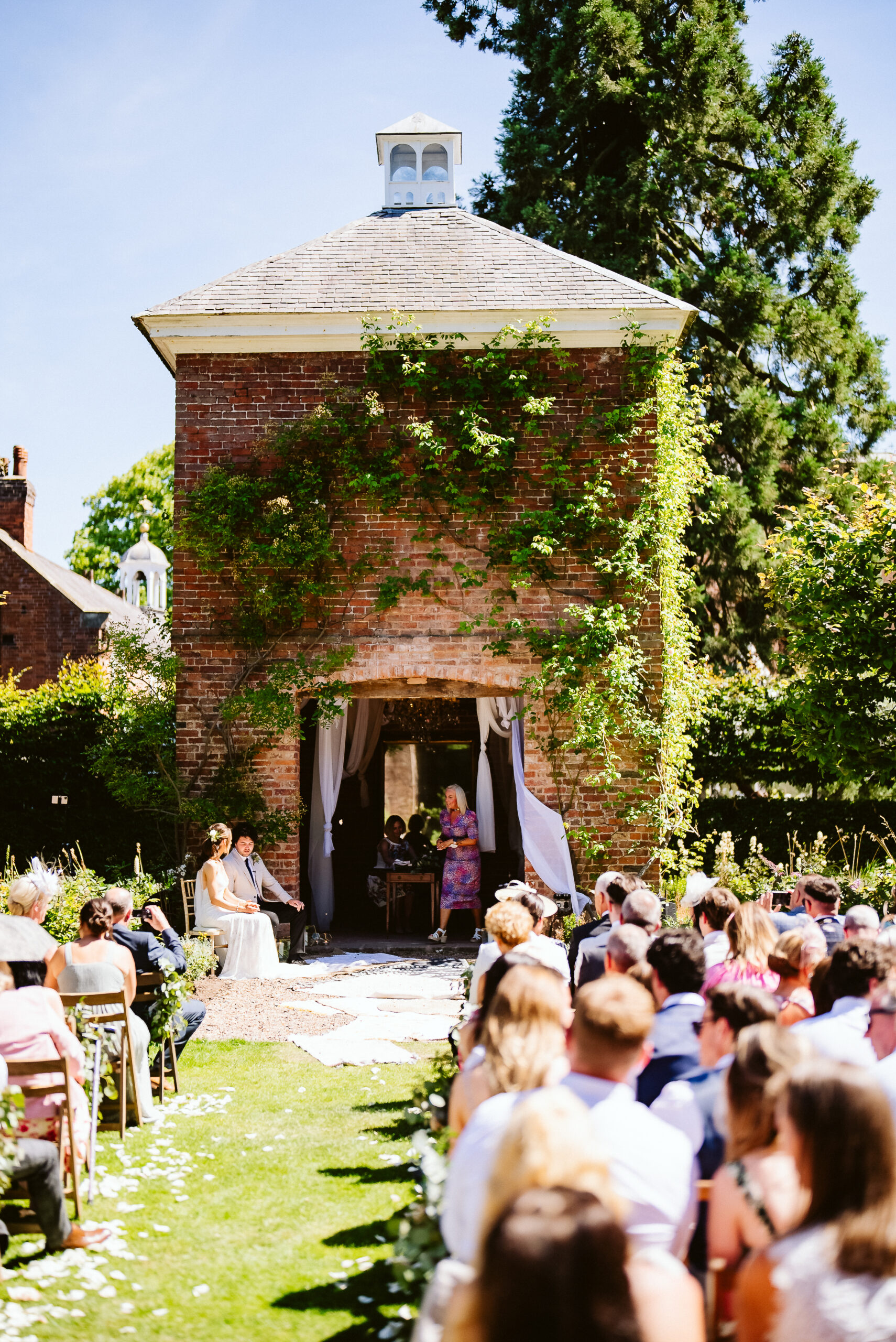 A tall redbrick square outhouse with a slate roof and climbing roses up the walls is in the background of the image and rows of brown wooden chairs have been set up with an aisle in between for an outdoor wedding ceremony