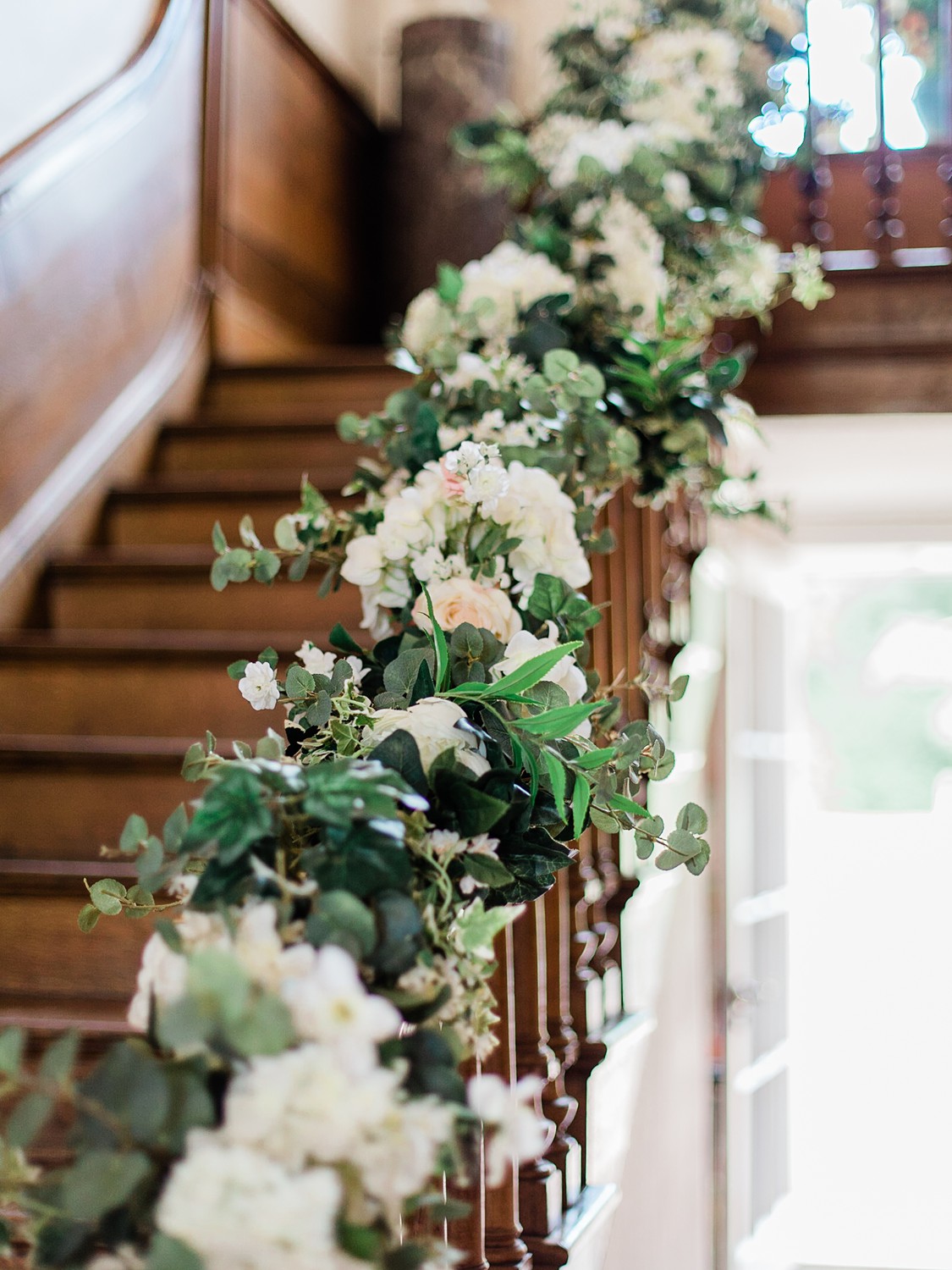 Roses, hydrangeas and white blossoms mixed with ivy and eucalyptus are entwined around the bannister of a wooden Georgian bannister