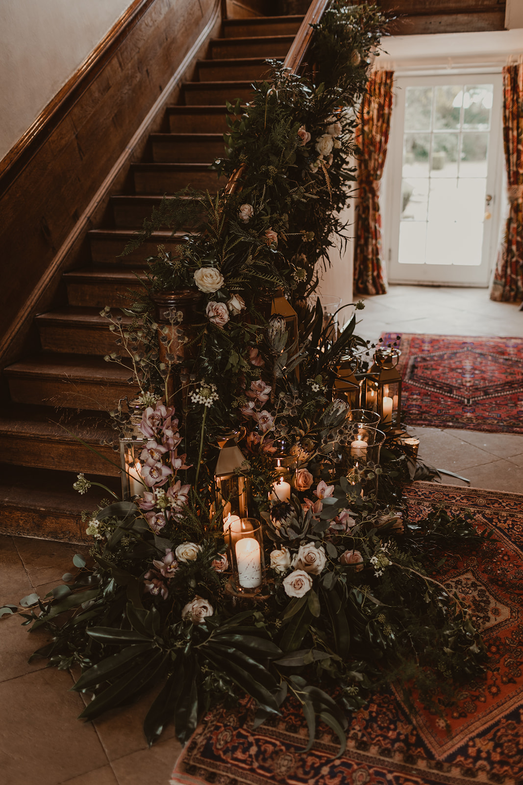 View of the side of an antique wooden staircase that has candles arranged at the bottom and soft pink flowers entwined around the bannister