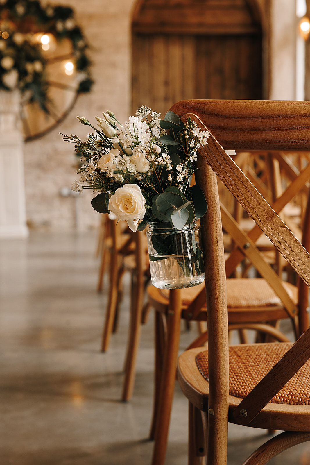 A close up view of a pew end hanging from the end of a wooden chair consisting of a jam jar filled with white roses, wax flower and lisianthus