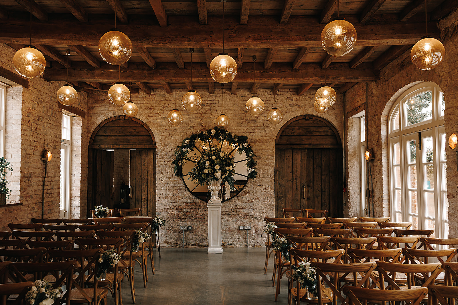 Rows of wooden chairs are lined up with spacing for an aisle to run between them inside a rustic converted coach house with exposed brick walls snd globe lights hanging from the ceiling