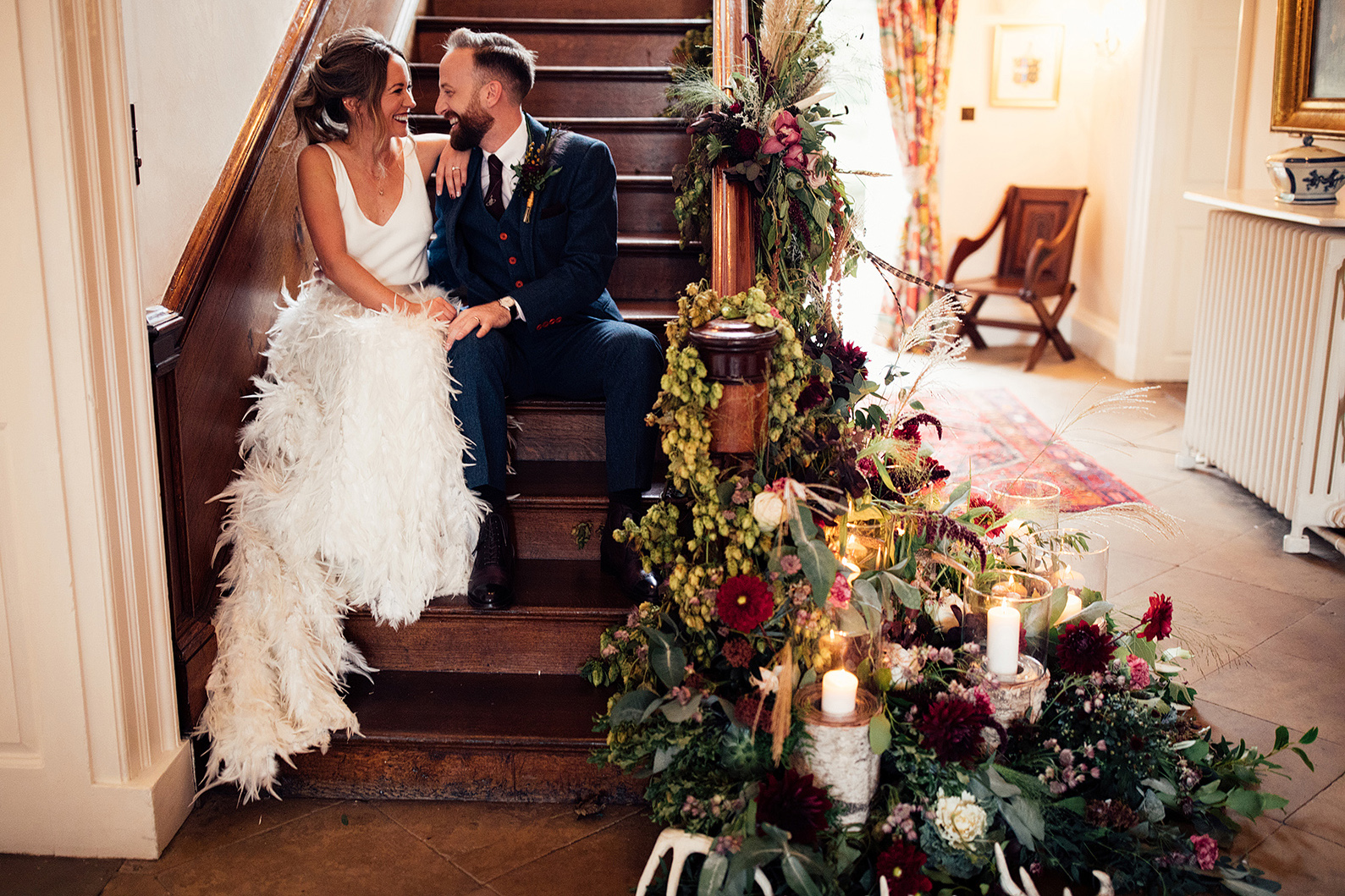 A bride and groom sit at the bottom of a set of antique wooden stairs surrounded by candles and Autumnal foliage
