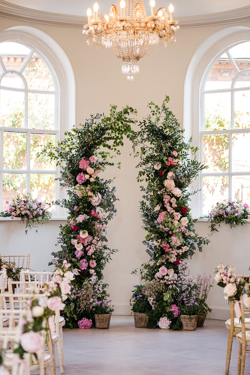 Two very tall pillars of foliage and pink florals are situated at the end of a large room that is flanked by two arched windows