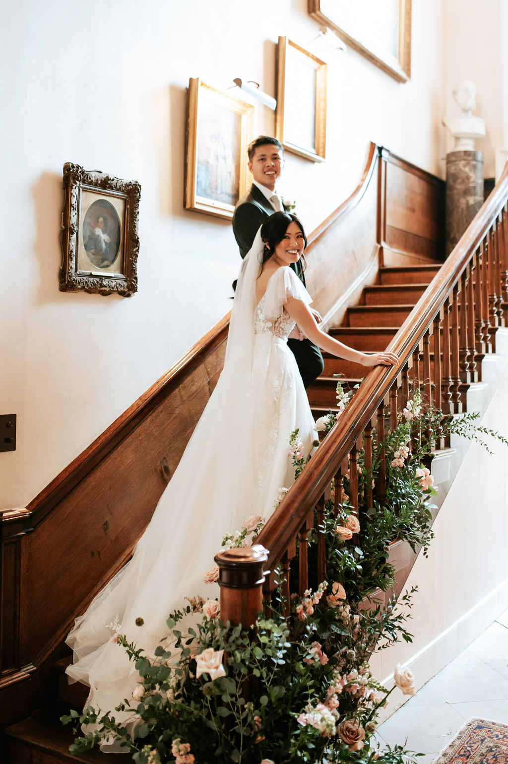 A bride in a white wedding dress and long veil walks up an antique wooden staircase that has candles arranged at the bottom and white and green flowers entwined around the bannister